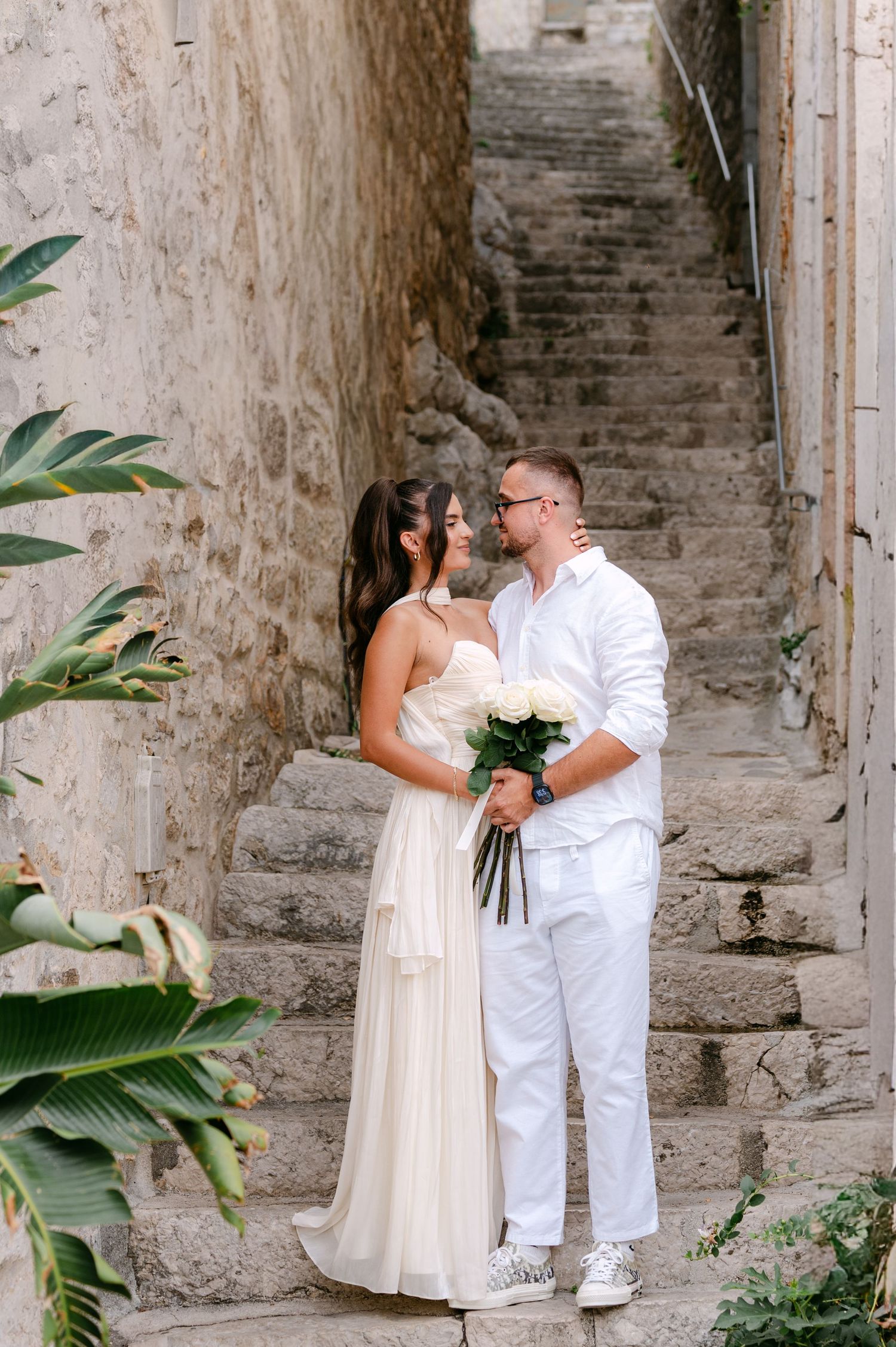 Couple in white wedding attire share a romantic moment on ancient stone staircase in old town.