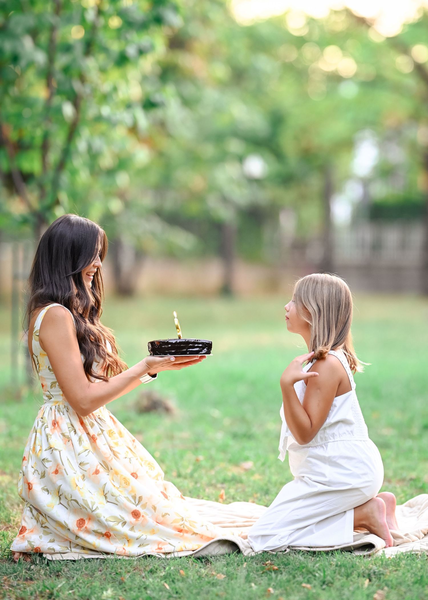 Mother presenting lit birthday cake to young daughter sitting on grass in garden setting.