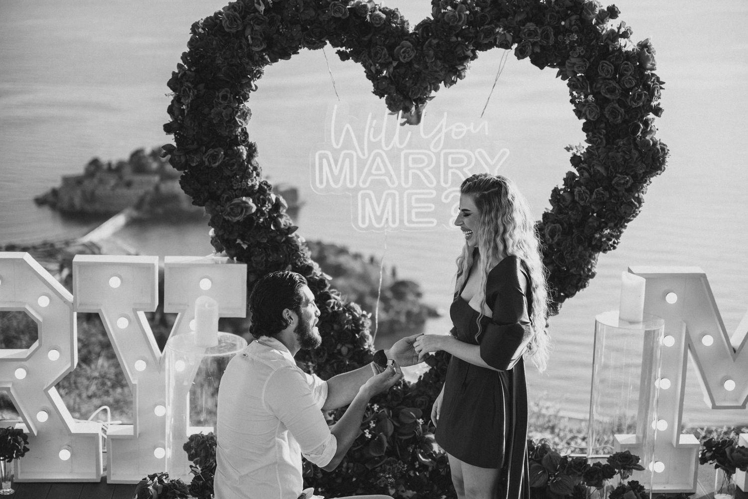 Black and white photo of man on one knee proposing to woman under a floral heart arch with 'Marry Me' sign.