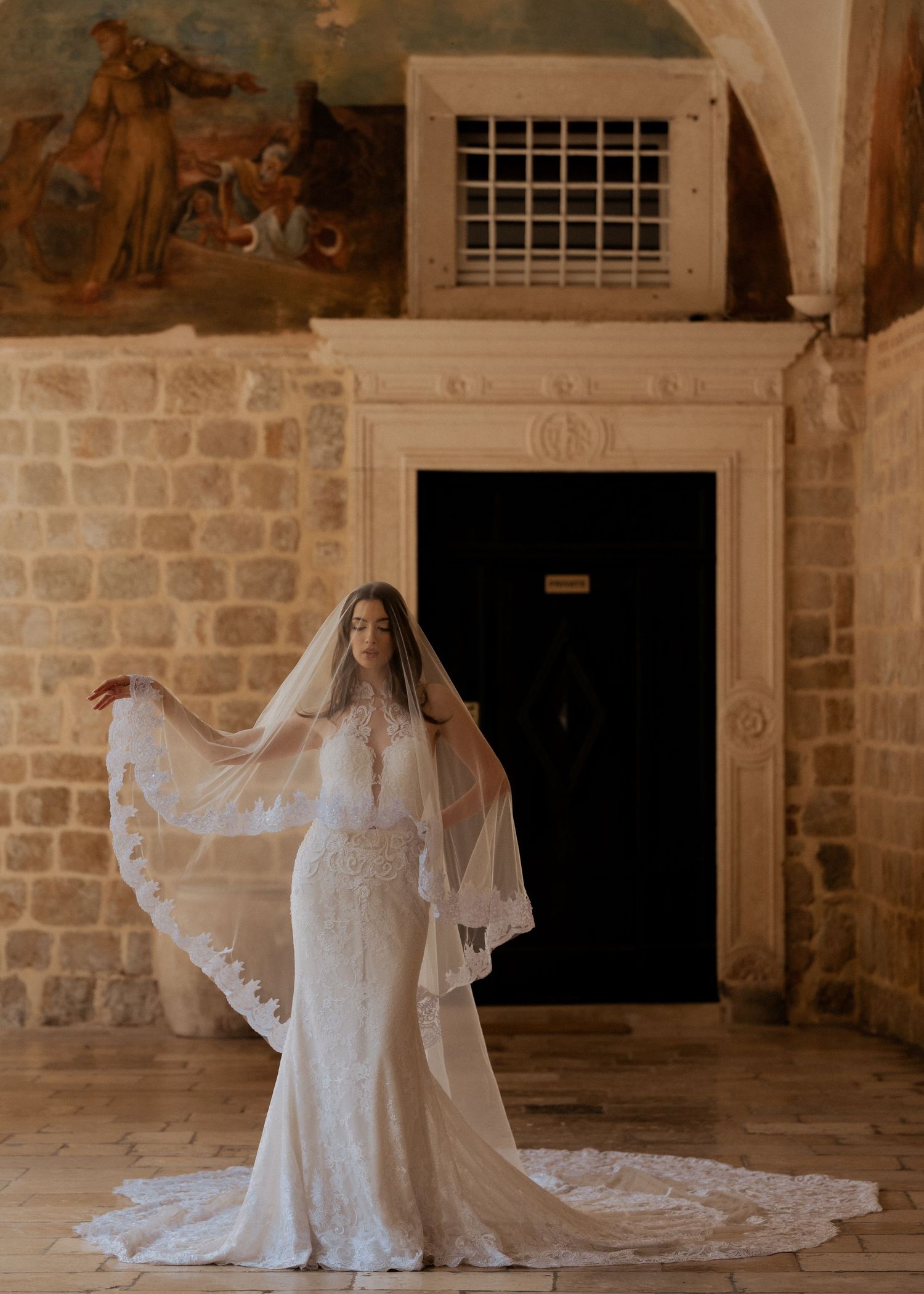 Bride in lace wedding gown with cathedral veil spreads arms in historic stone chapel with ornate frescoed ceiling.