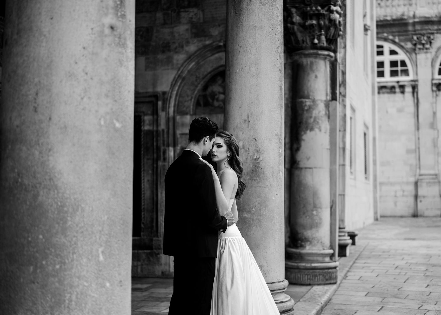 Bride and groom embracing romantically between grand stone columns in a classic black and white photo.
