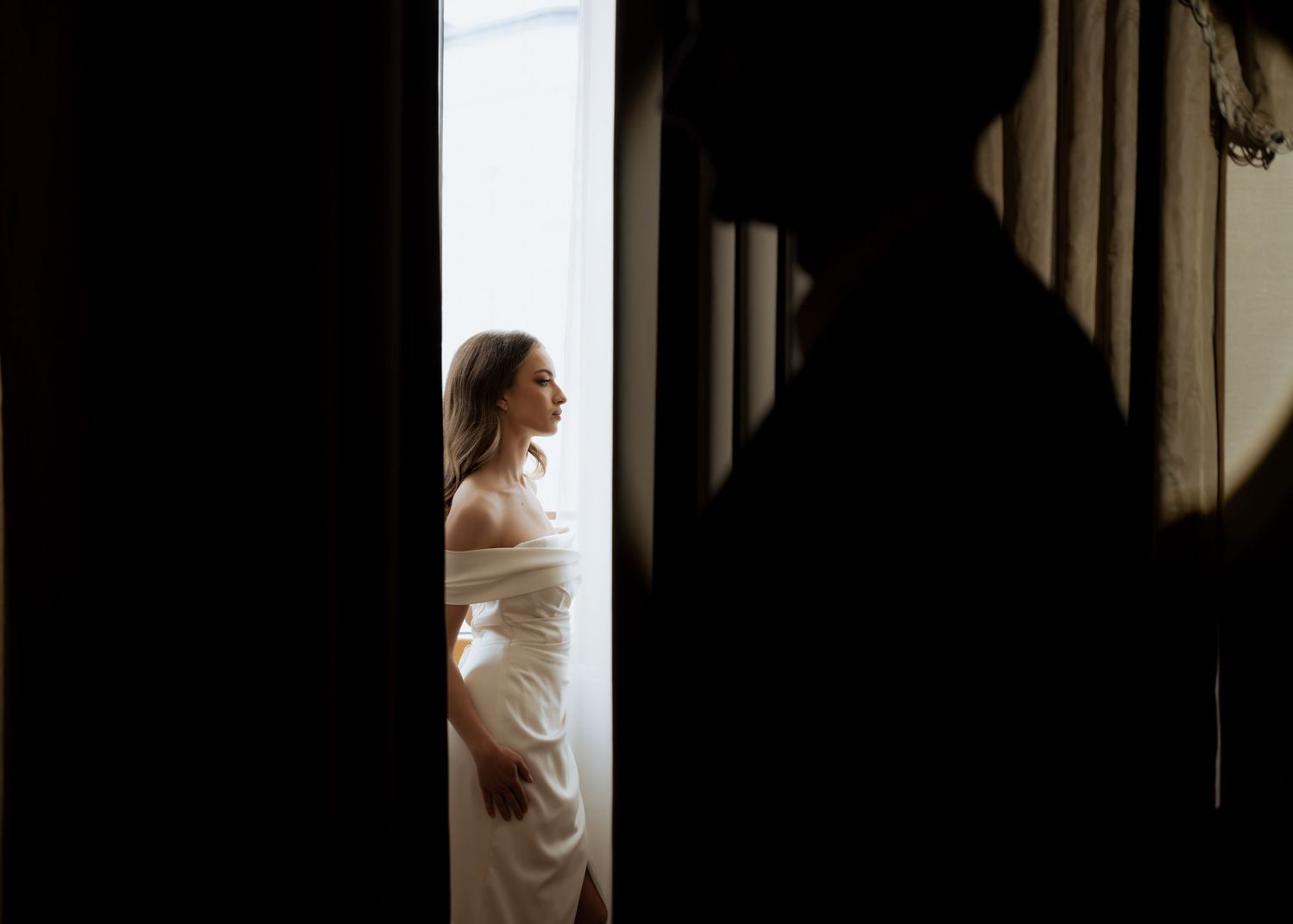 Dramatic doorway shot of bride in white gown illuminated by light while groom stands in shadow.
