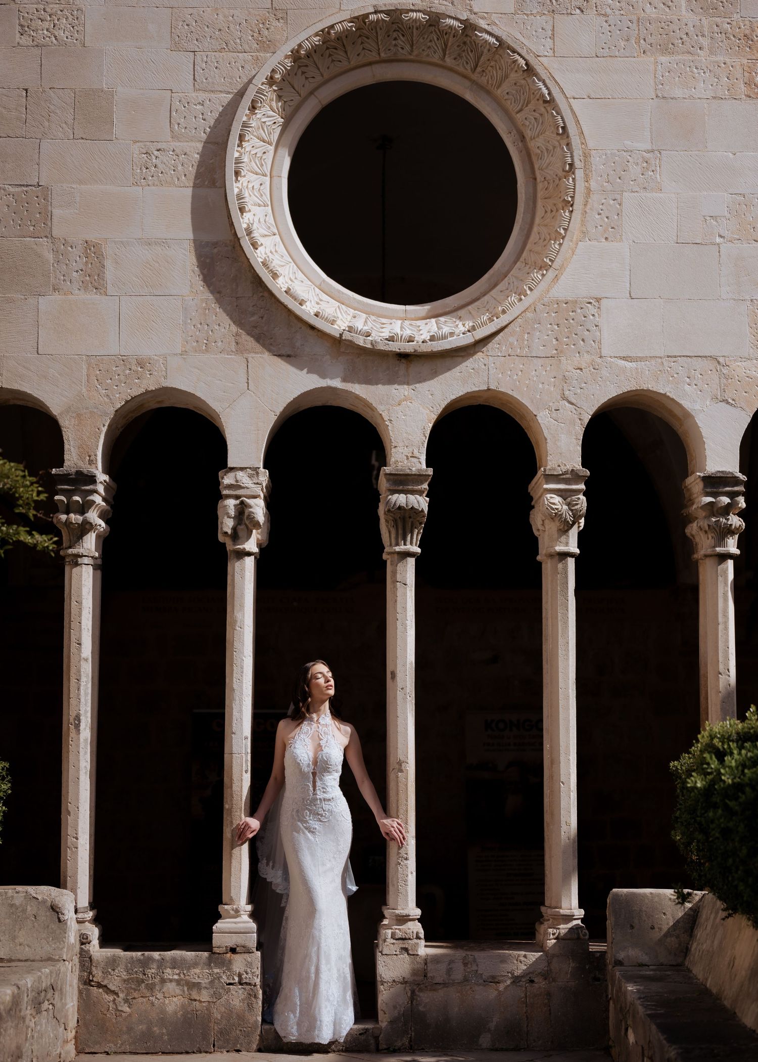 Bride in a fitted white gown standing beneath ornate stone arches and a circular window of a historic building.