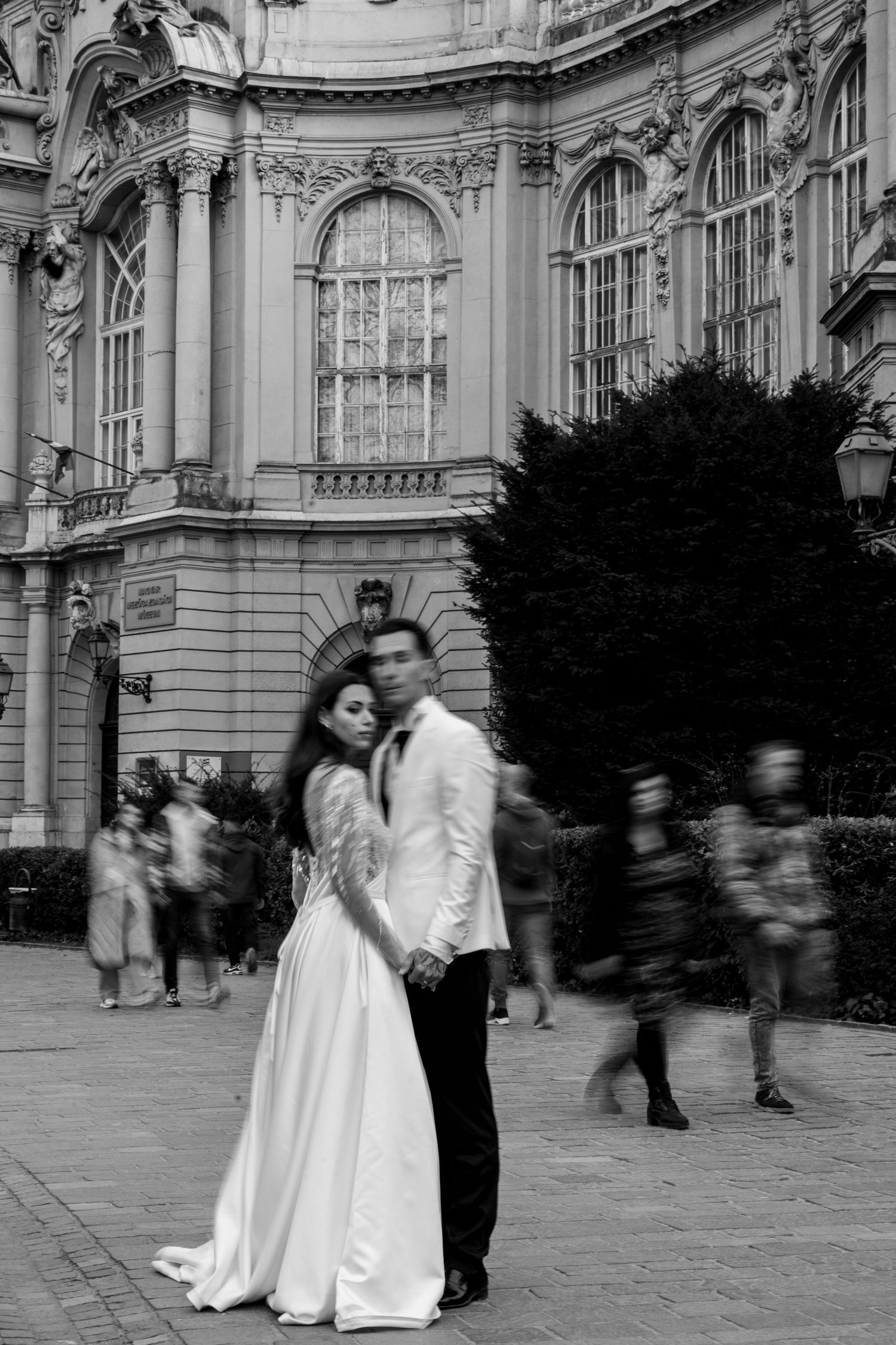 Bride and groom embracing in black and white photo with blurred crowd and ornate European building.