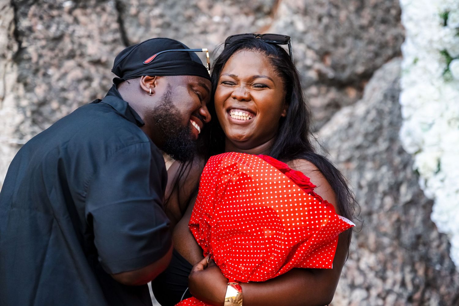 A couple shares a joyful moment, the man whispering to a laughing woman in a red outfit.