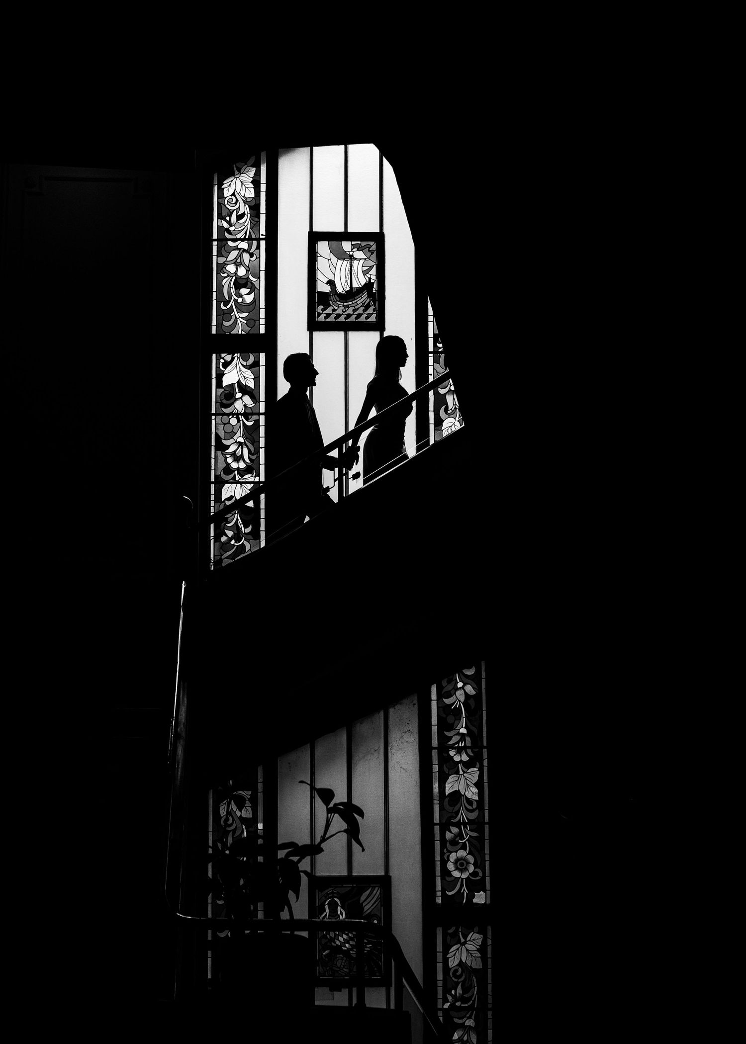 Silhouette of bride and groom on spiral staircase beside ornate stained glass window in black and white.