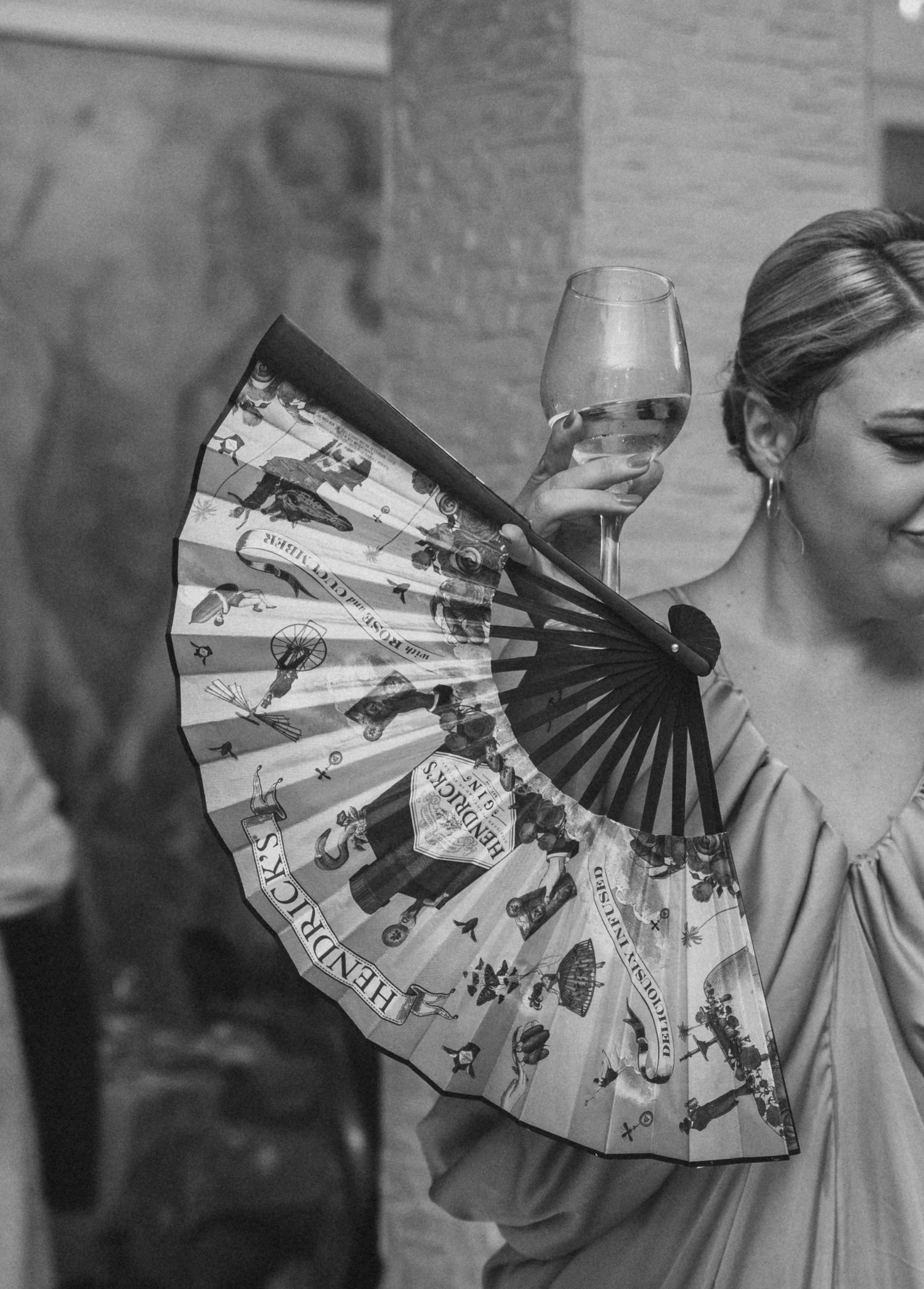 Black and white photo of woman holding decorative fan and wine glass at elegant event.