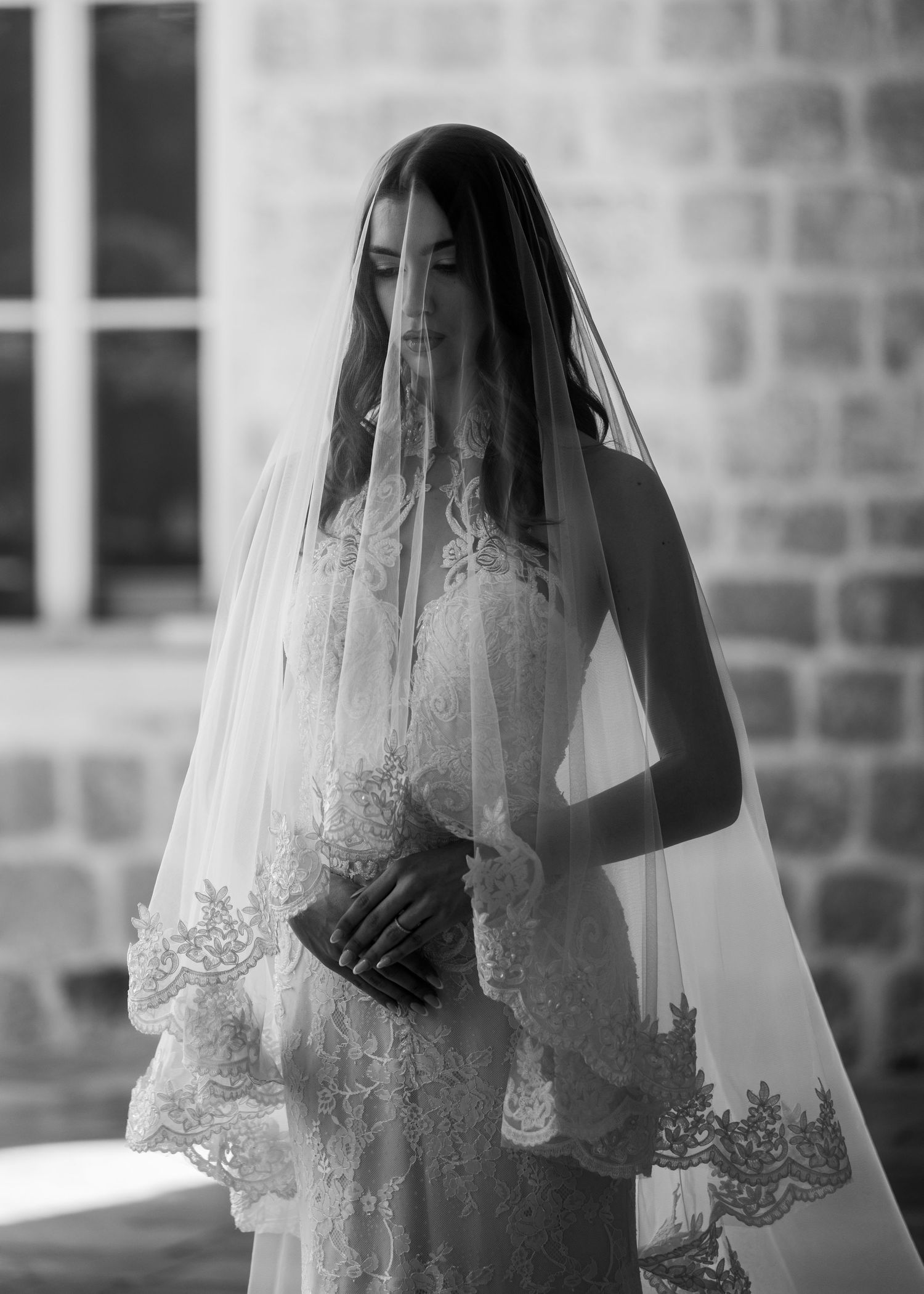 Black and white portrait of a bride wearing a lace-trimmed veil over her face in soft light.