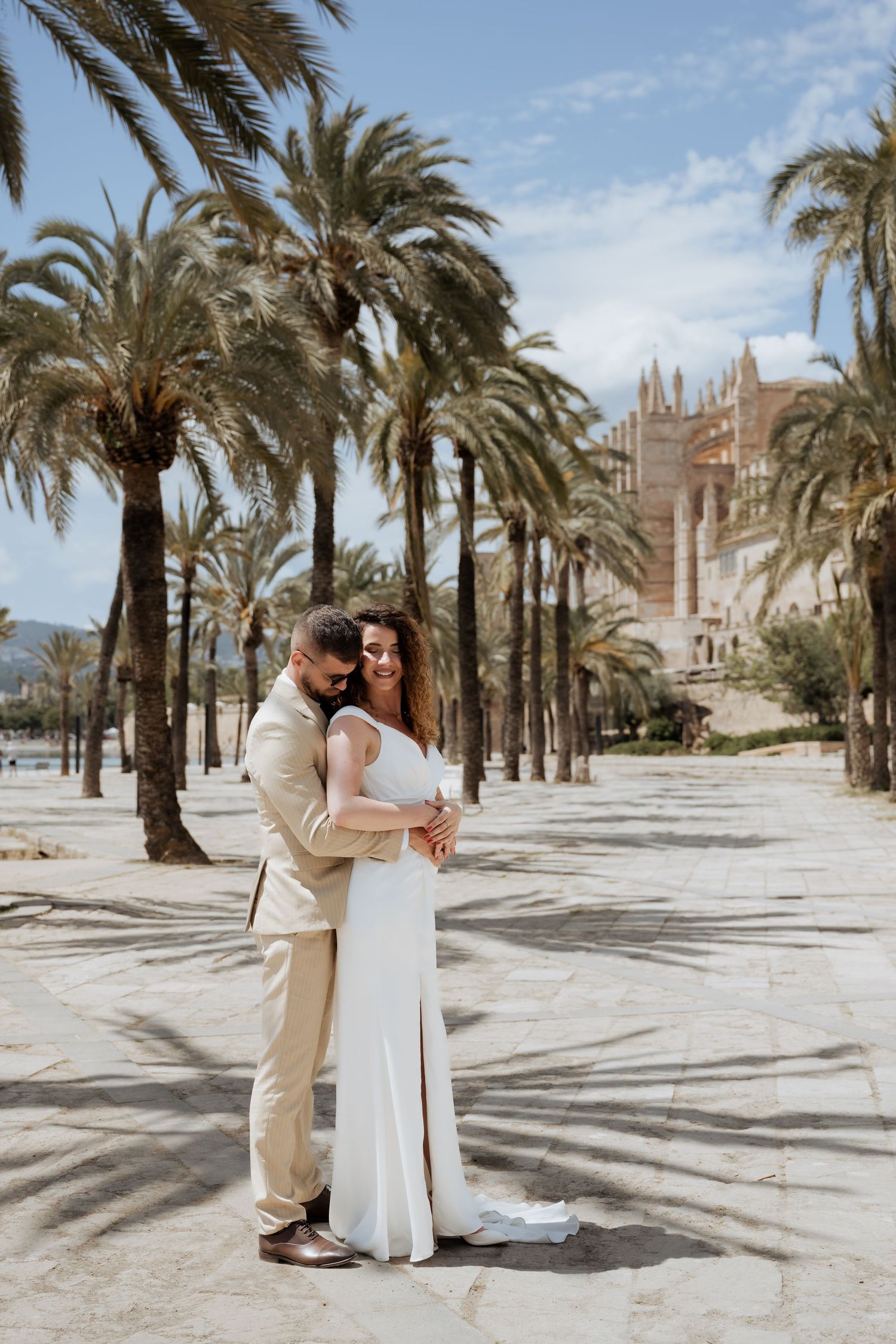 Groom embraces bride from behind as they pose among palm trees near Gothic cathedral.