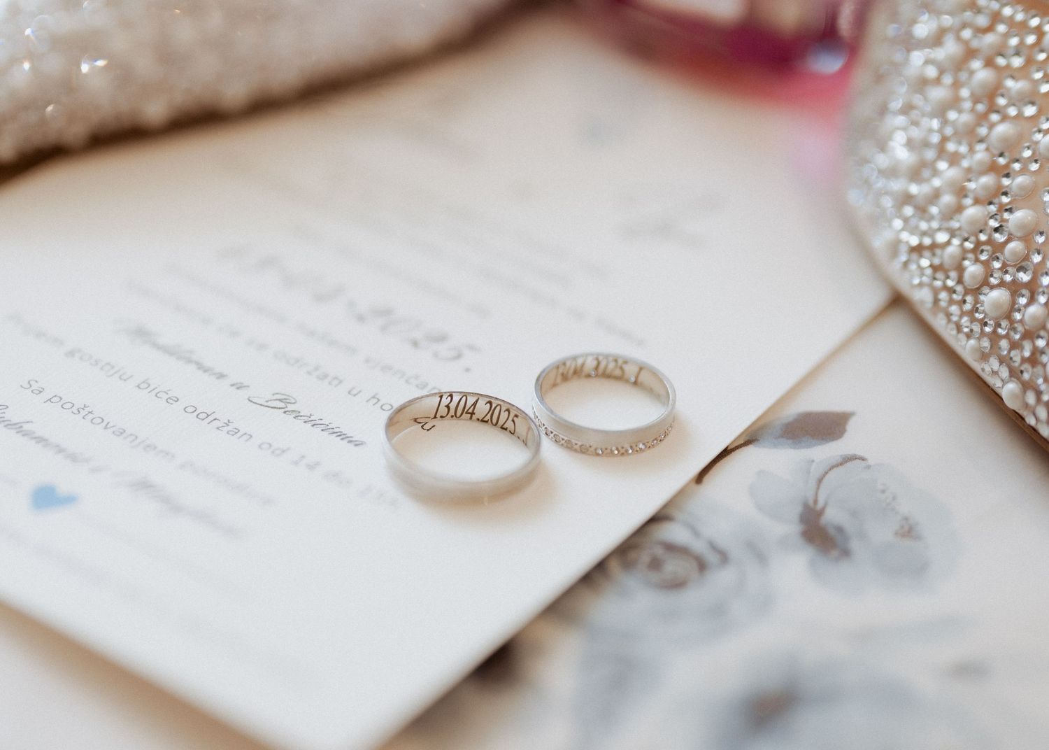 Two engraved wedding bands resting on wedding invitation with pearl-embellished fabric nearby.
