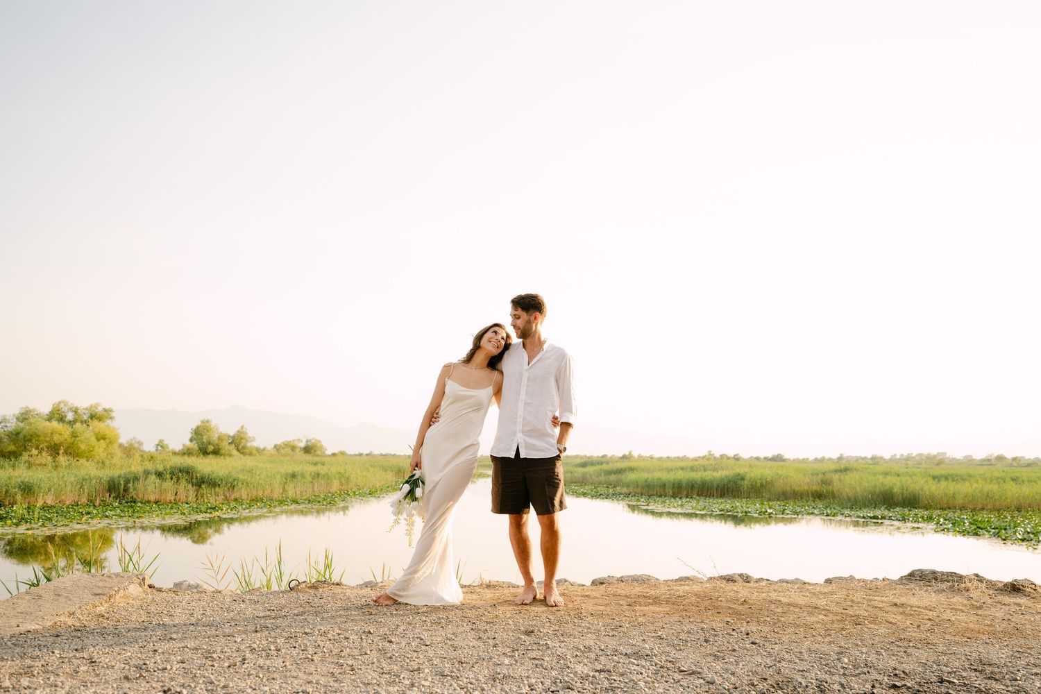 Bride and groom in white attire walking hand in hand near a serene lake at golden hour.