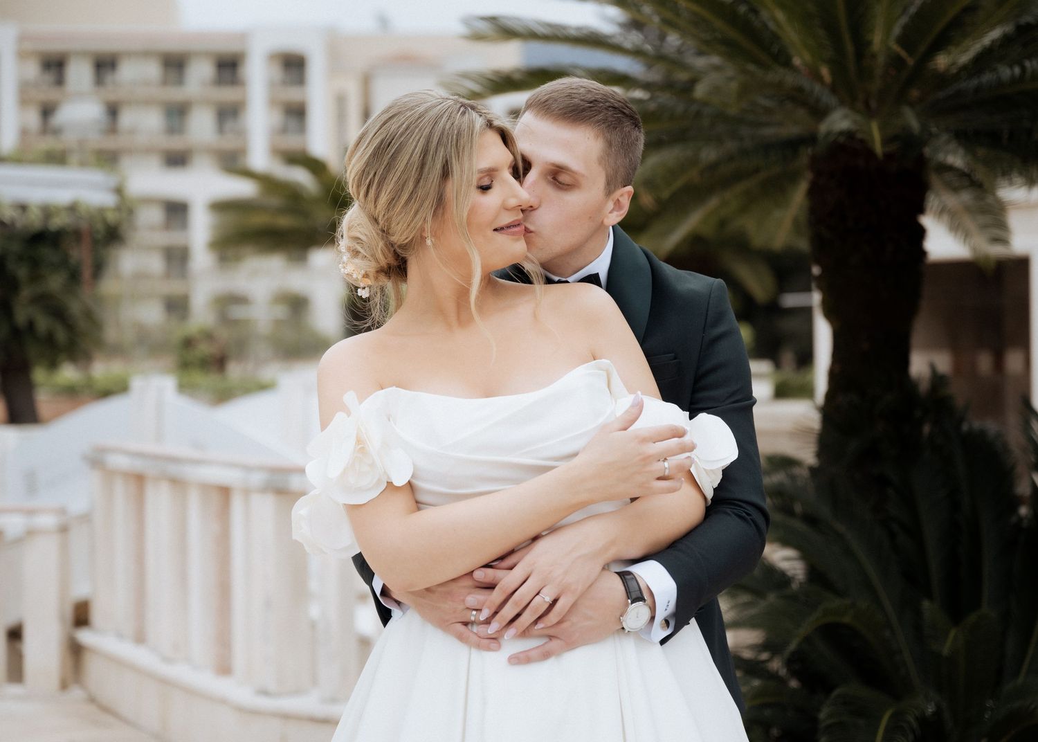 Romantic bride and groom embracing and kissing outdoors with palm trees in background.