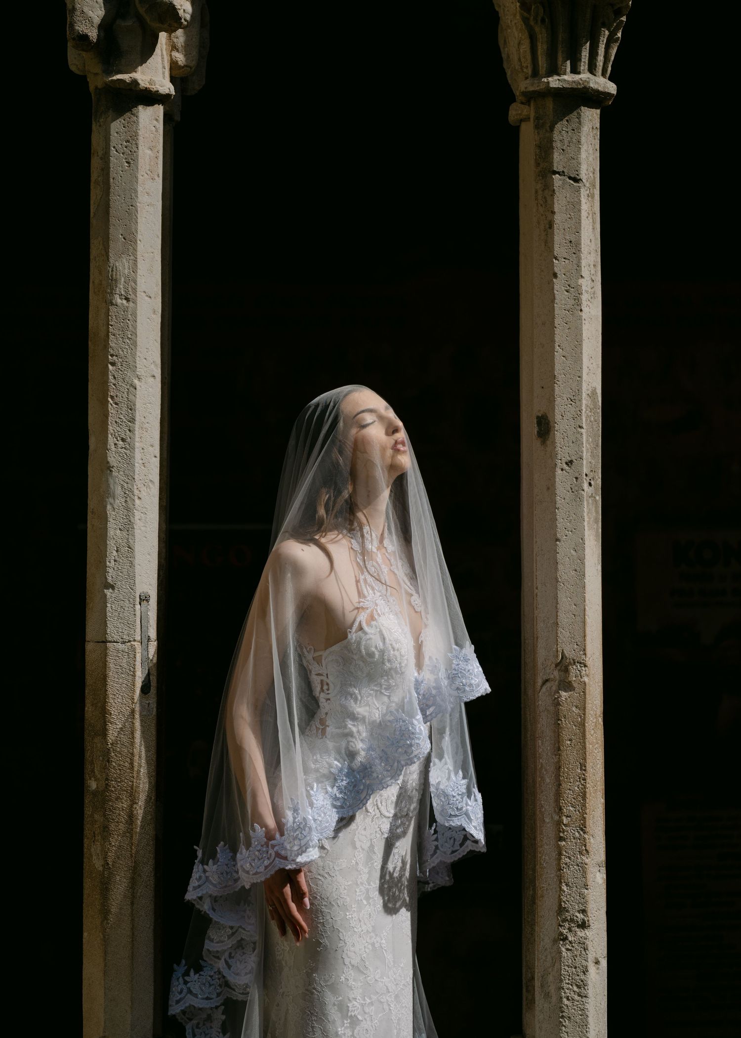 Ethereal bride in lace wedding gown and veil stands between ancient stone columns in dramatic light.