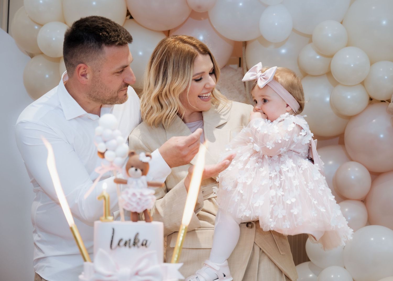 Parents with their baby girl celebrating first birthday named Lenka in front of balloon backdrop.