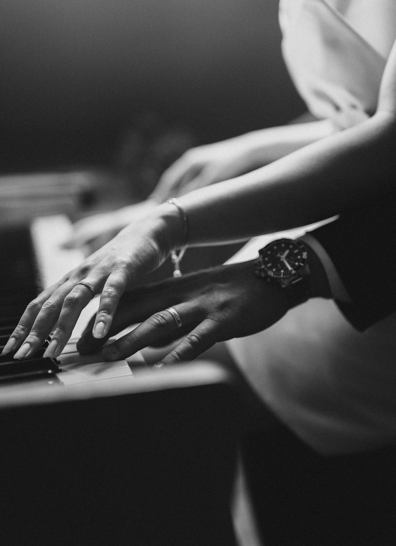 Close-up black and white photo of hands playing piano with watch and bracelet visible.