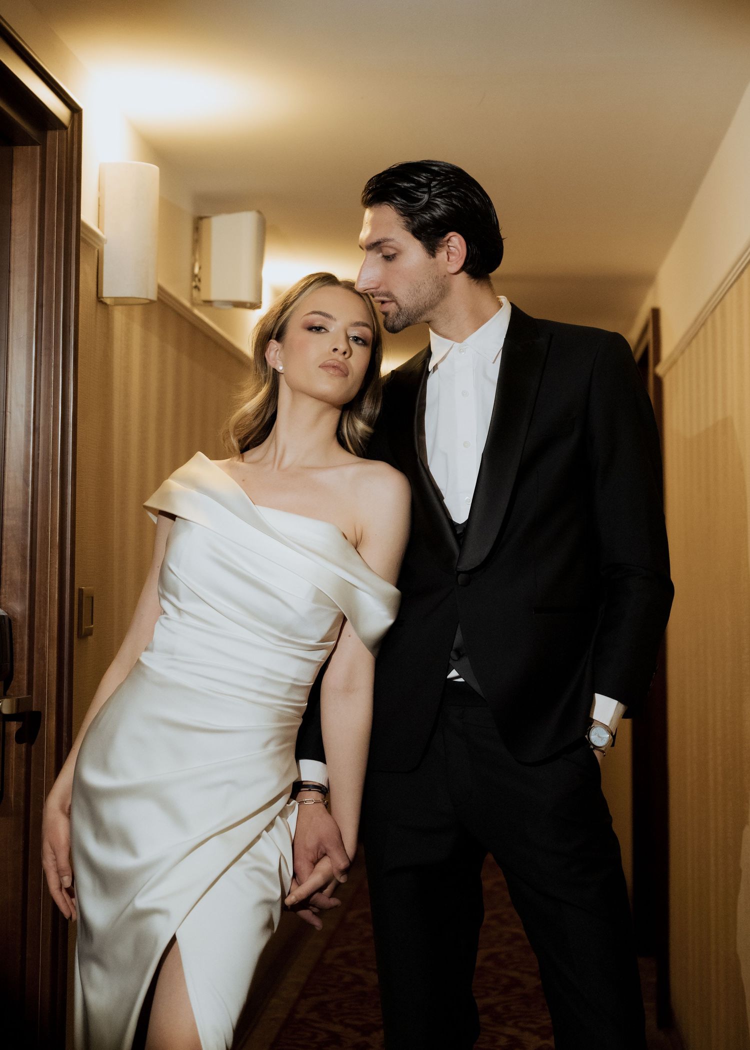 Stylish couple in wedding attire hold hands in warmly lit hotel corridor.
