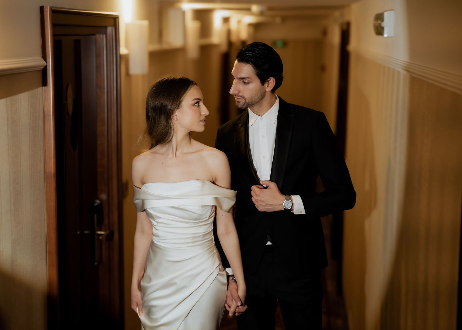 Elegant wedding couple holding hands in a hallway, bride in off-shoulder white gown, groom in black tuxedo.
