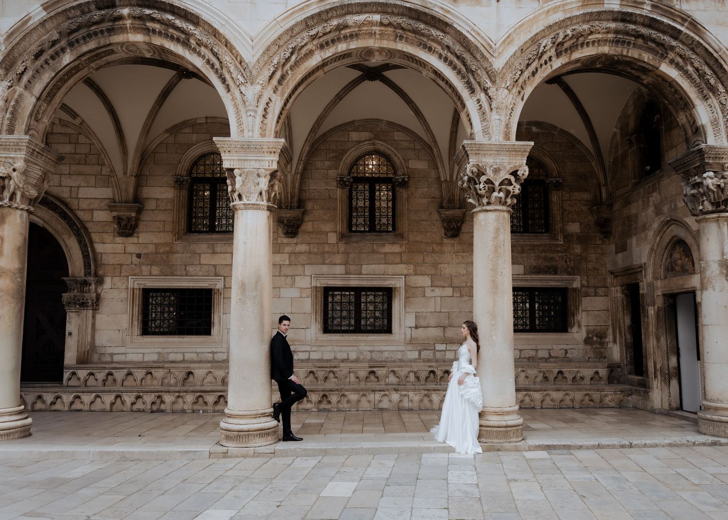Wedding couple standing apart in a grand stone courtyard with Gothic arched colonnades.