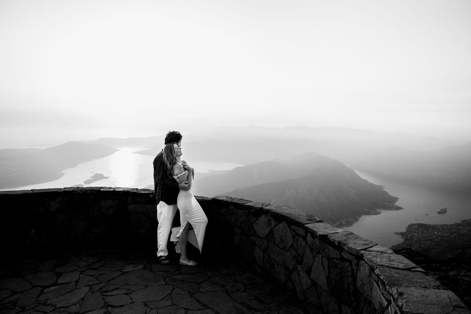 Black and white photo of a couple embracing at a scenic mountain overlook with a river valley below.