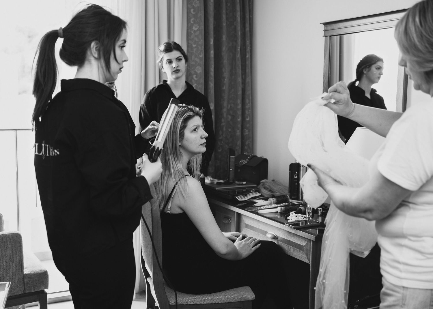 Black and white image of a bride seated at a vanity getting hair and makeup done by stylists.