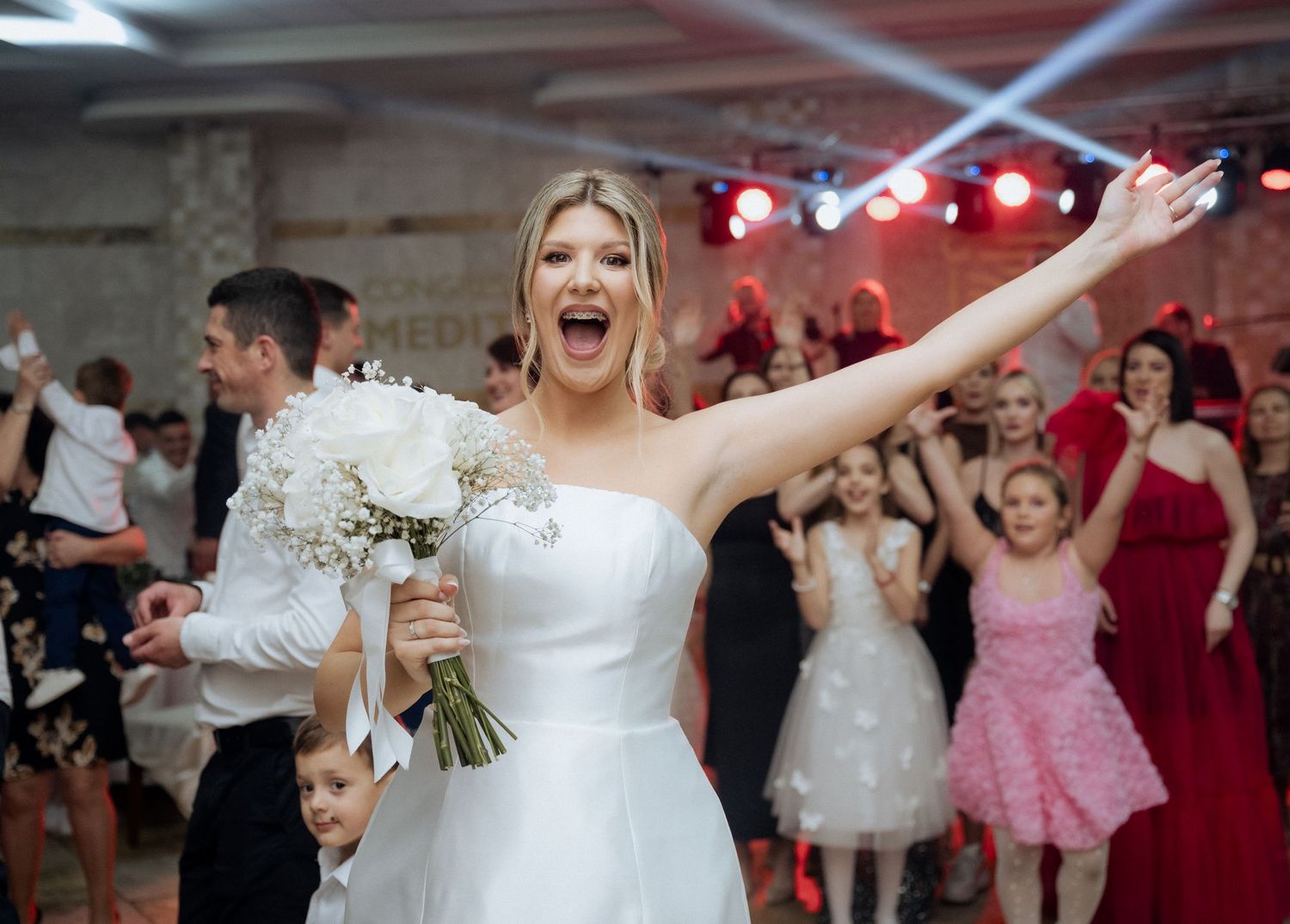 Joyful bride in white dress holding bouquet with arm raised at lively wedding reception.
