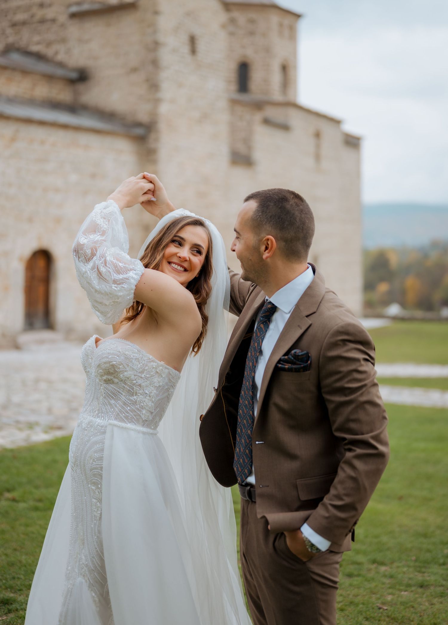 Bride and groom dancing outdoors near a stone church, bride in lace wedding dress with veil, groom in brown suit.
