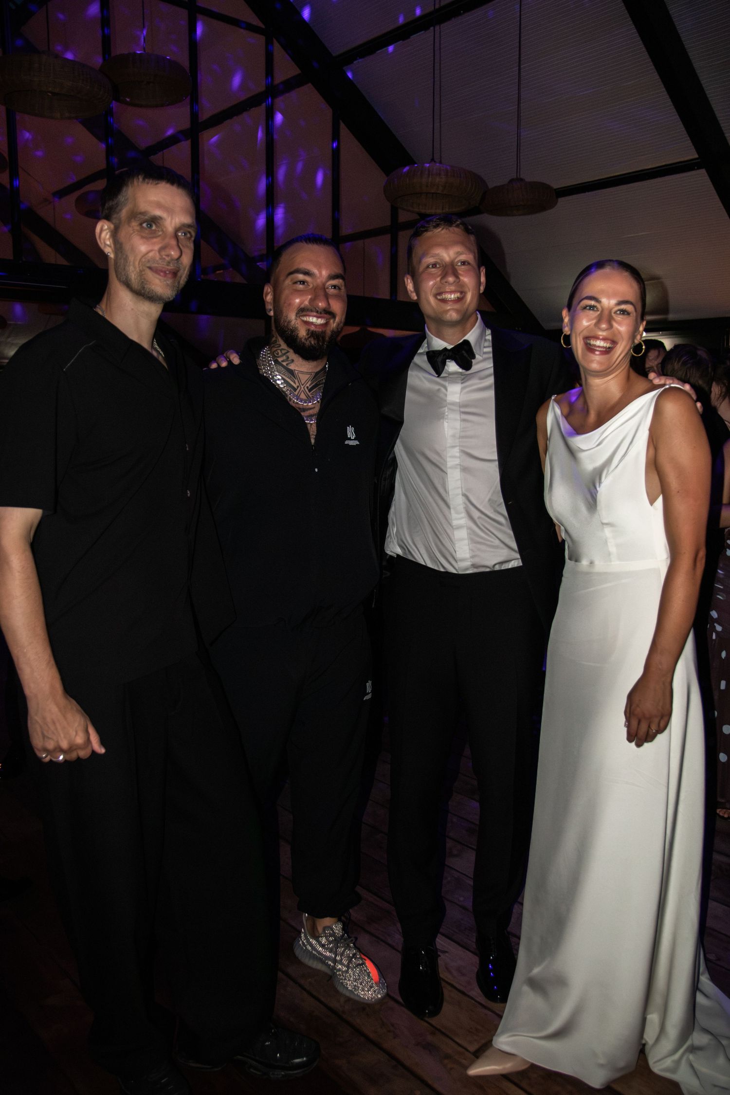 A bride in a white gown and a groom in a tuxedo pose with two smiling friends dressed in black at a lively evening wedding reception.