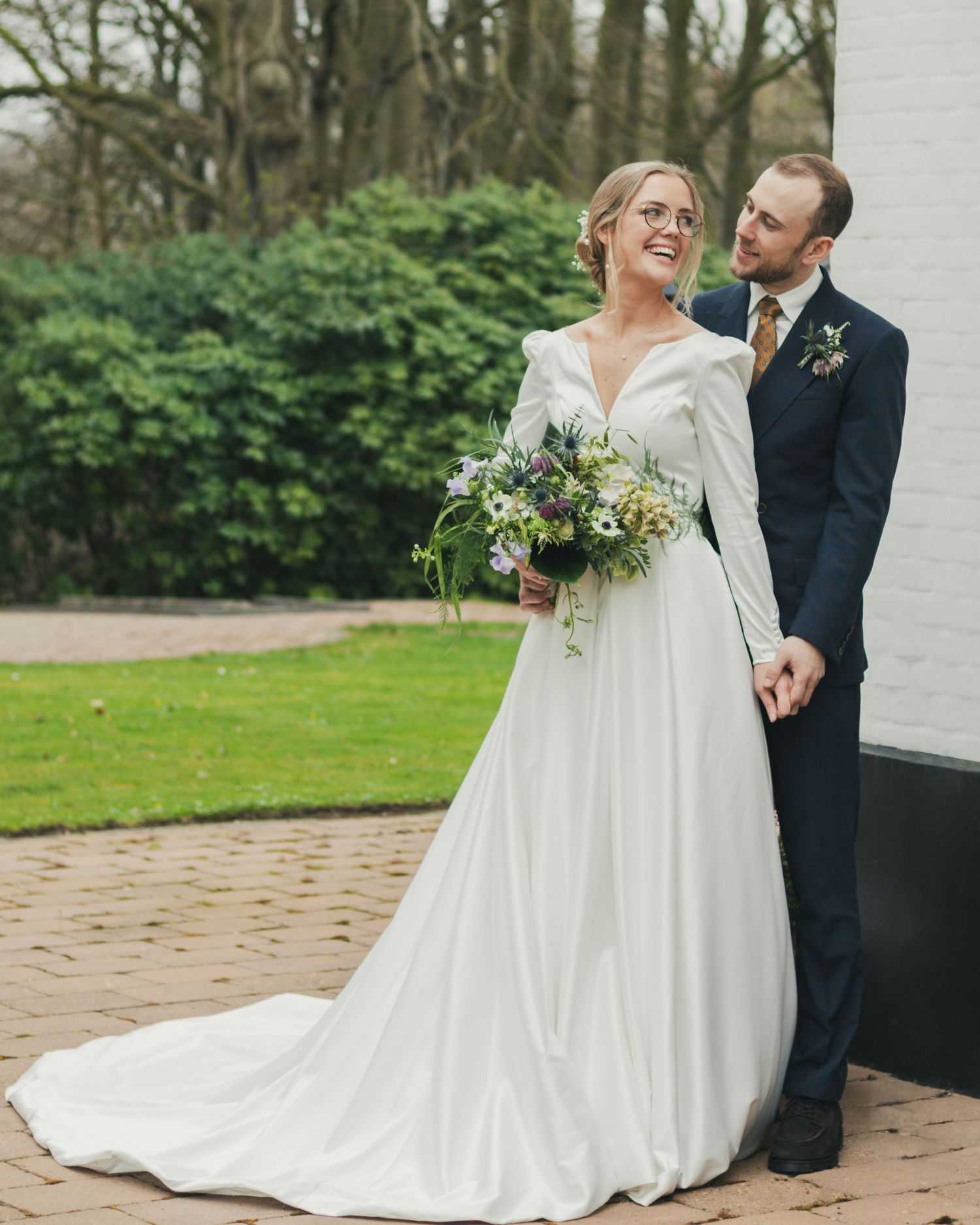 Bride in a long white gown holds a wildflower bouquet while her groom in a navy suit stands behind her on a brick patio.
