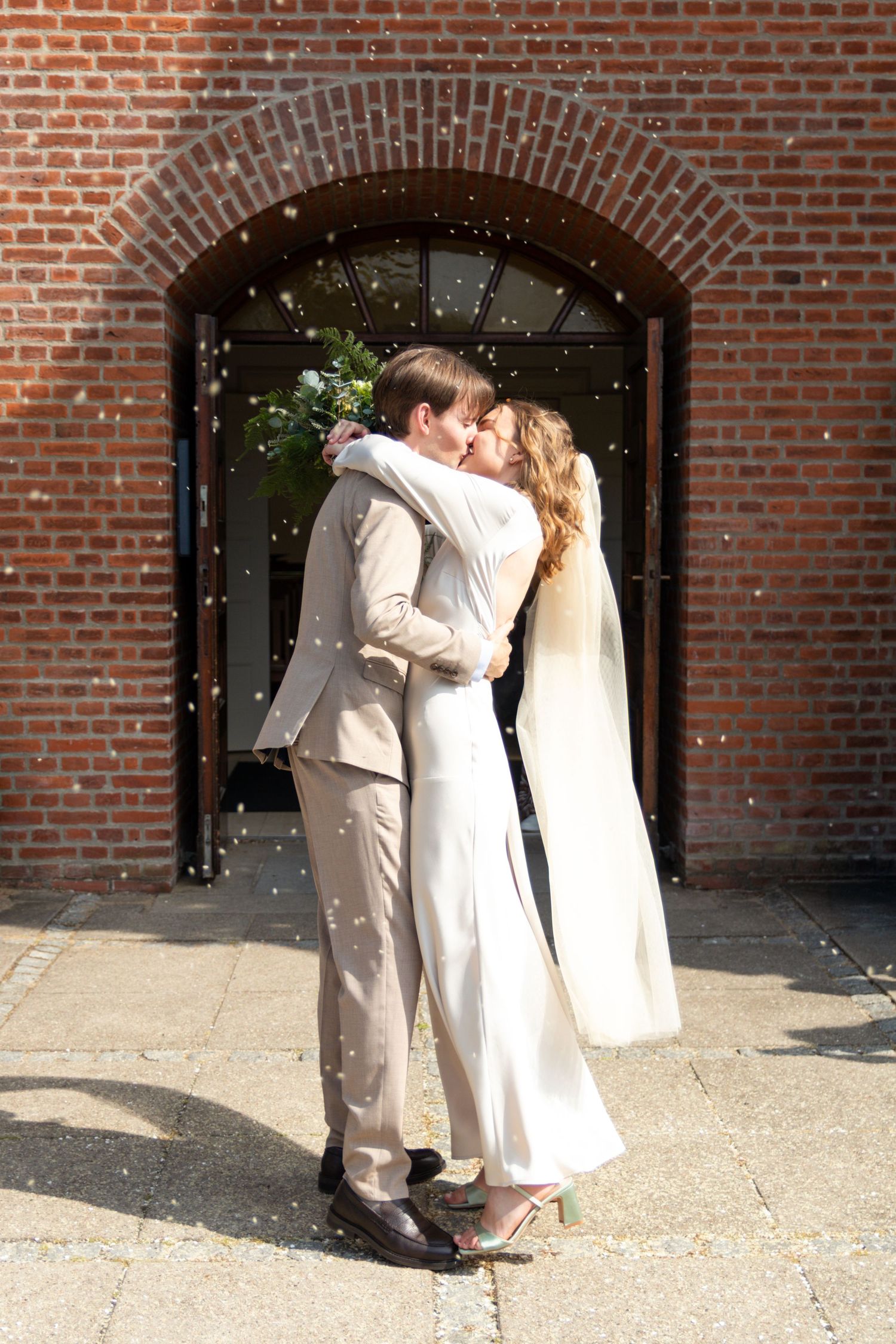 Groom in a beige suit and bride in a white dress share a kiss in front of a brick church doorway as confetti falls.