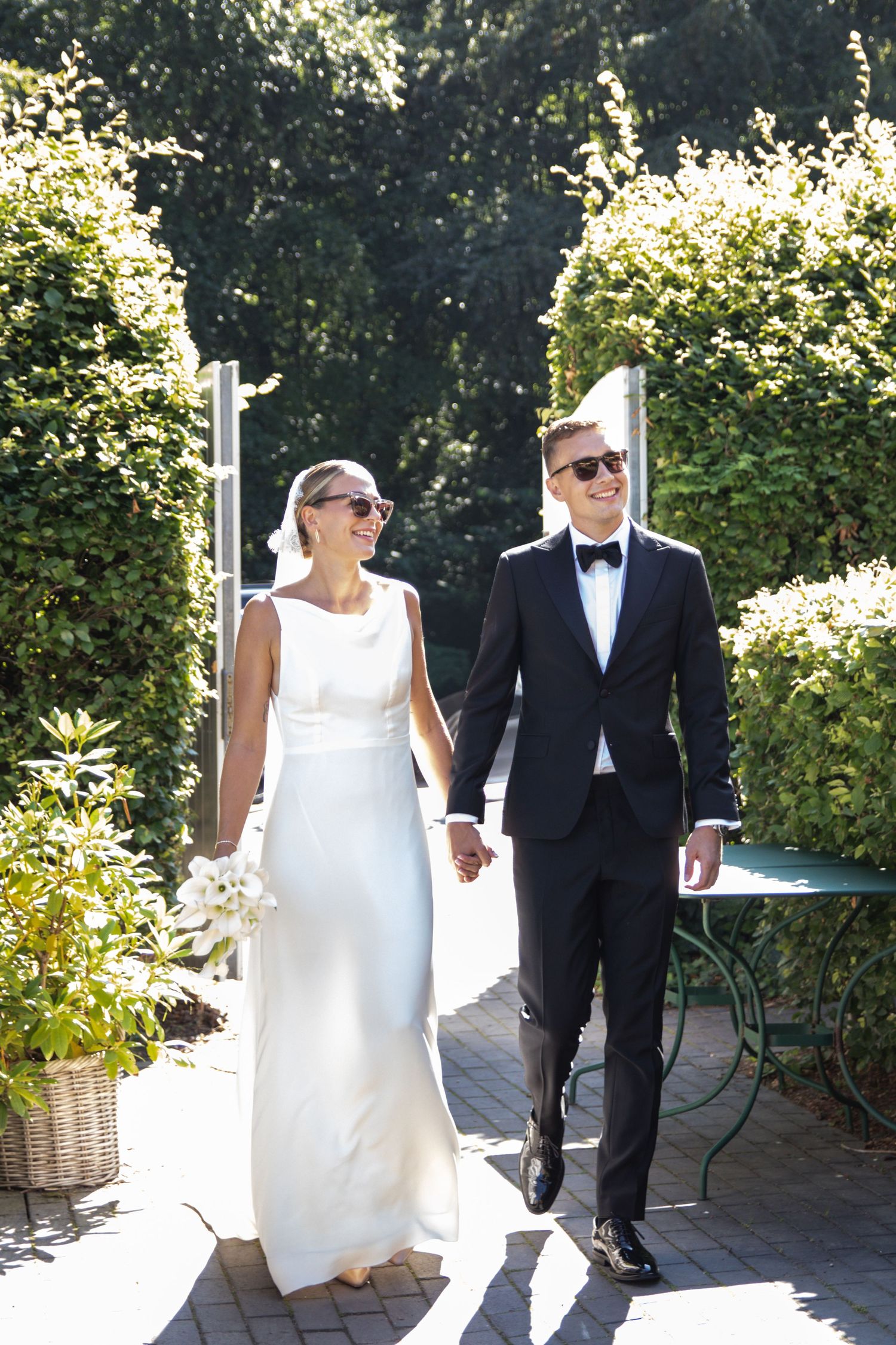 A smiling bride and groom in sunglasses walk hand in hand through a sunny garden path lined with green hedges.