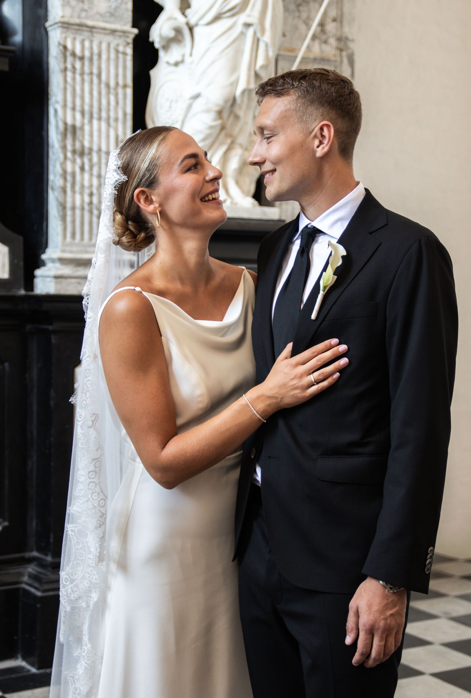 A smiling bride in a satin gown with a lace veil rests her hand on her groom's chest inside a grand marble hall.