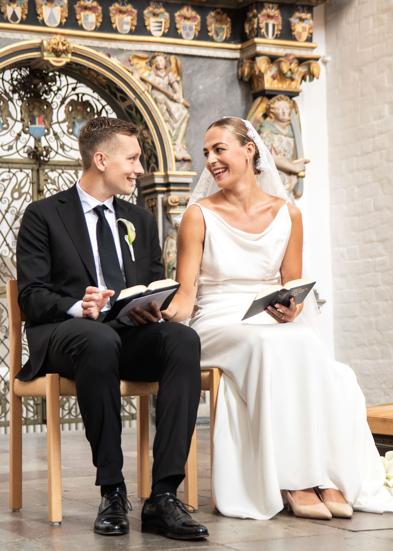 Smiling bride and groom sit side by side on wooden chairs in a historic church, holding wedding service books.