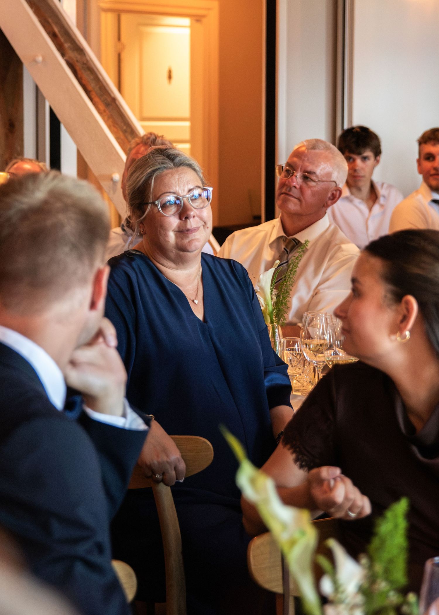 A woman in a navy dress and glasses and other wedding guests listen attentively to a speech at a reception table with flowers.