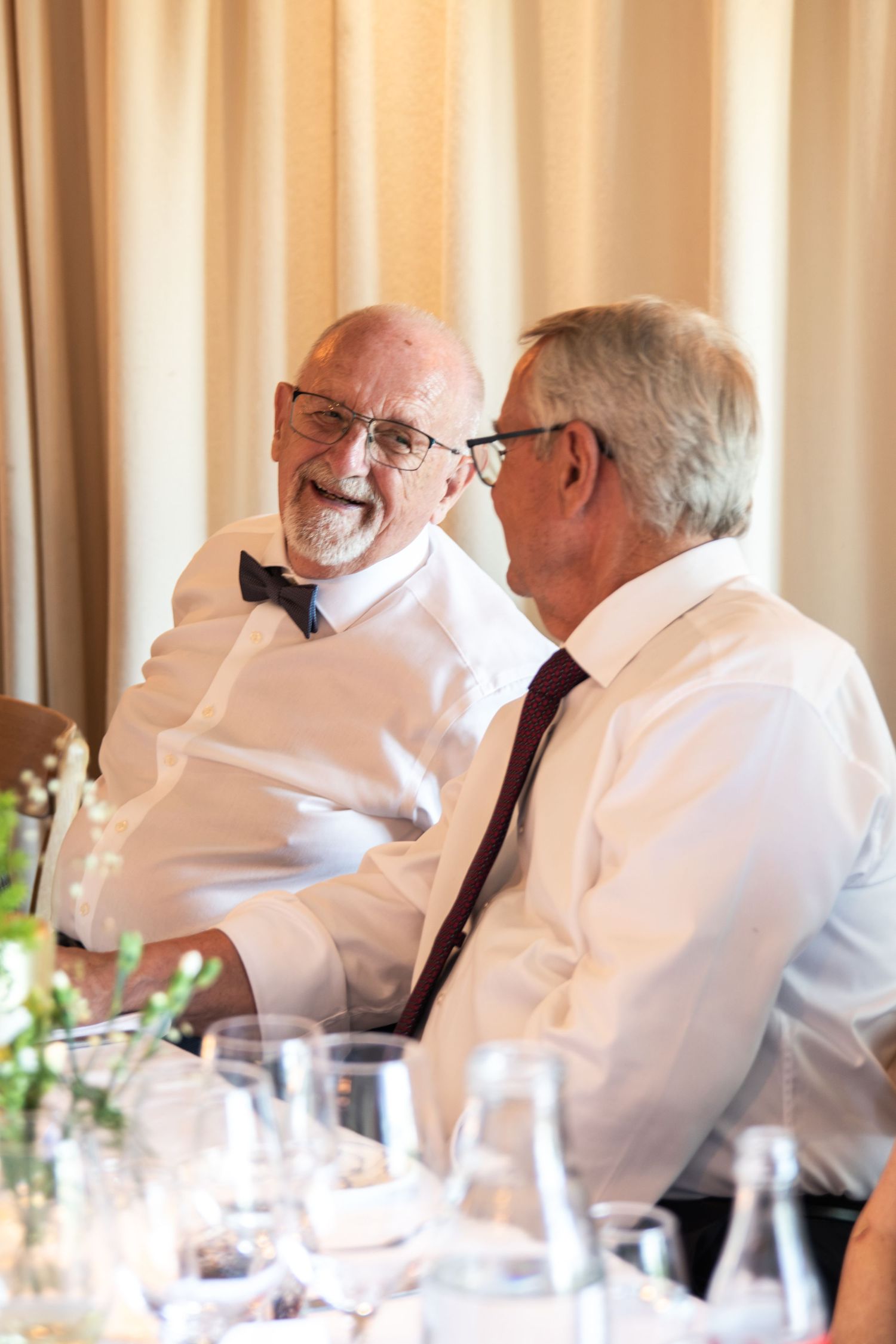 Two older men in white shirts laugh and chat together at a wedding reception table with glasses and fresh greenery.