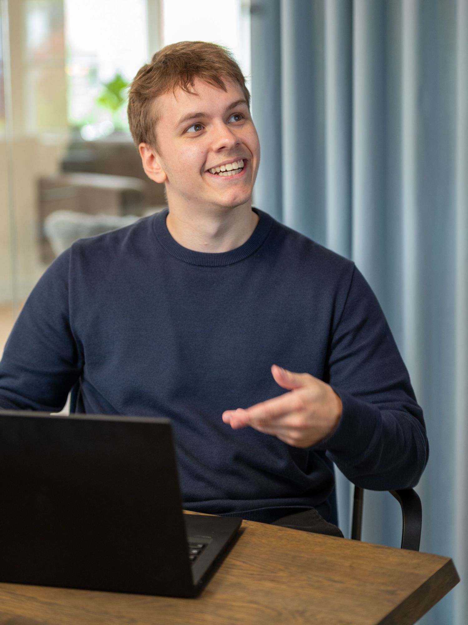 A young man in a navy sweater gestures and smiles while sitting at a wooden table with a laptop in a bright office.