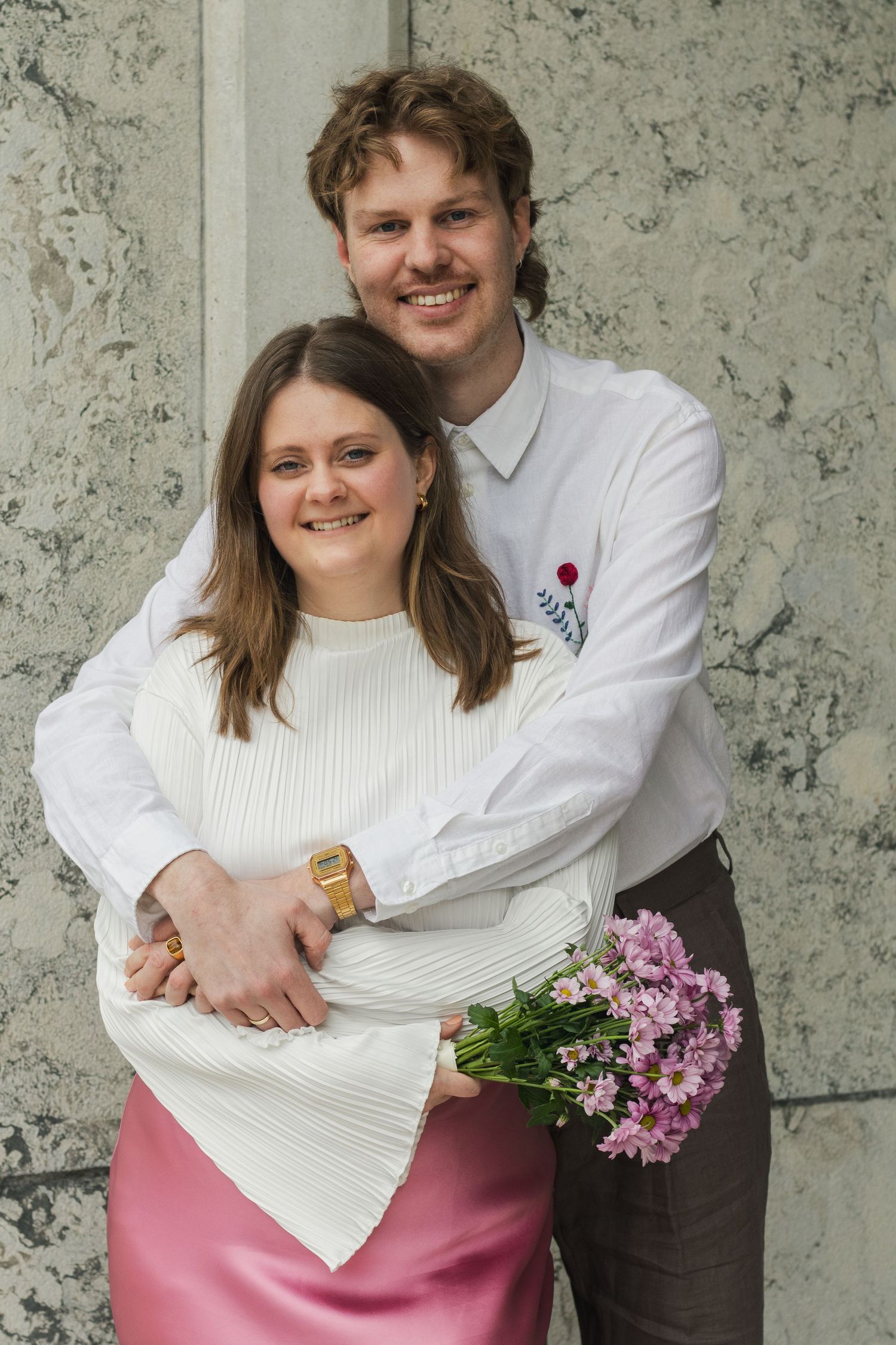A smiling man hugs his partner from behind as she holds a bouquet of pink flowers, both standing against a marble wall.