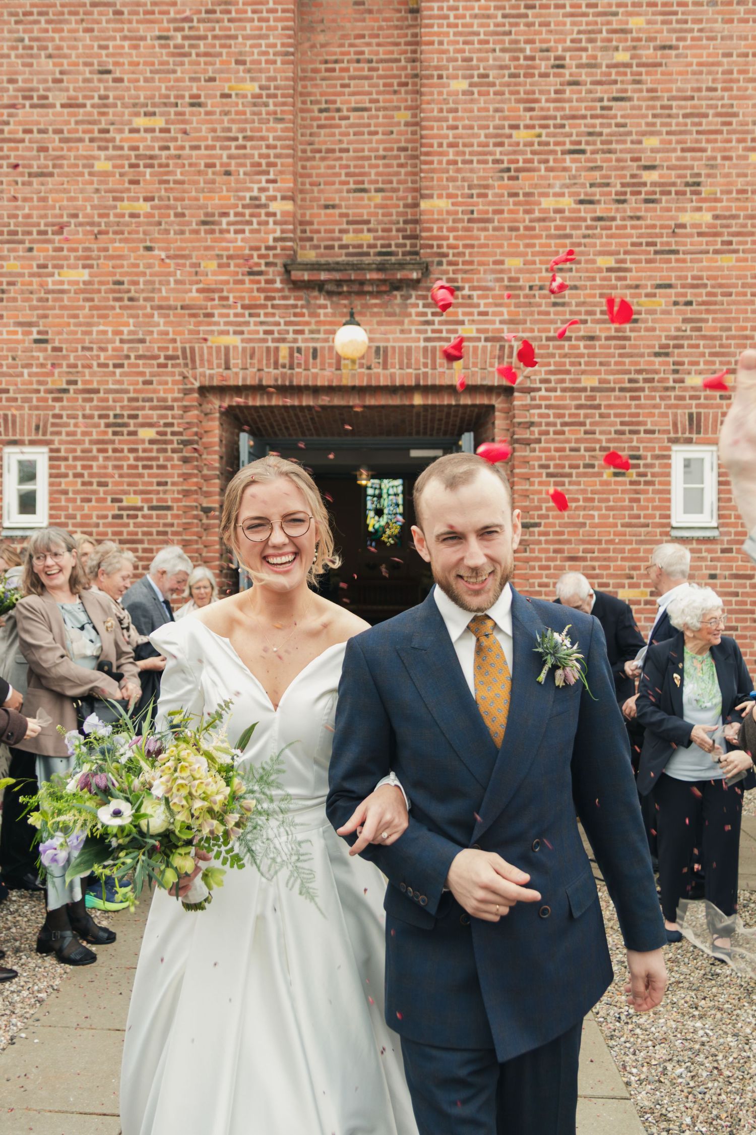A laughing bride and groom walk out of a brick church as guests toss red rose petals into the air around them.