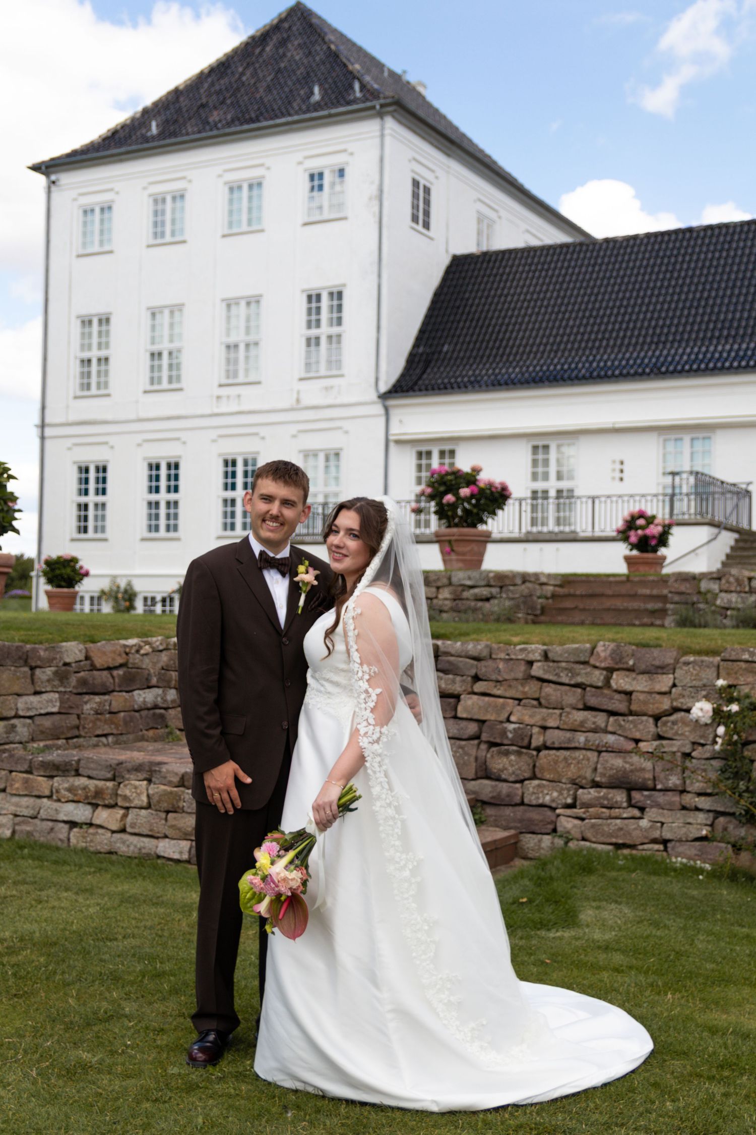 Bride in a white gown and groom in a brown suit pose on a lawn in front of a white manor house with a dark roof.
