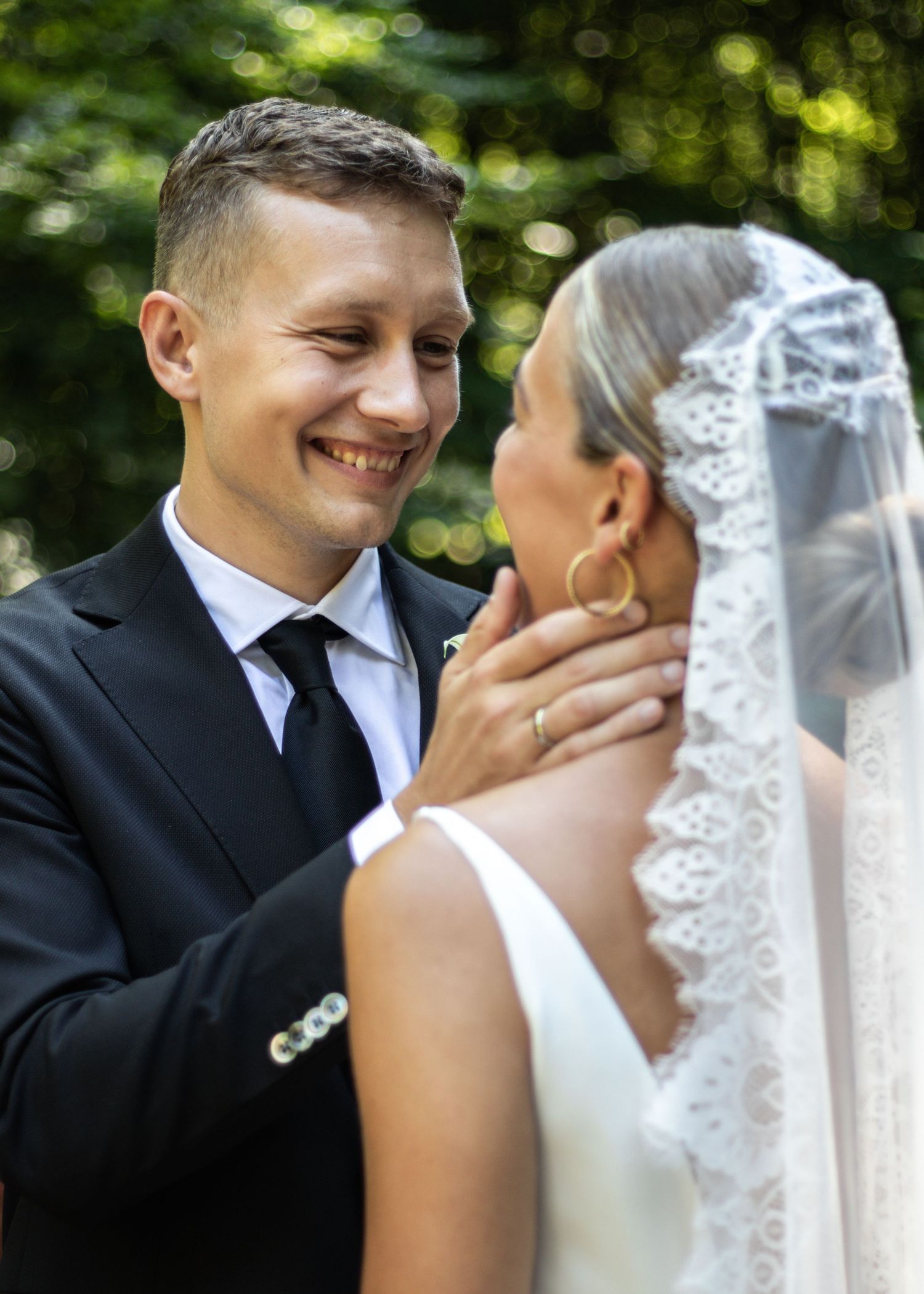 A close-up of a smiling groom in a black suit touching his bride's face as she wears a delicate lace veil and gold hoop earrings.