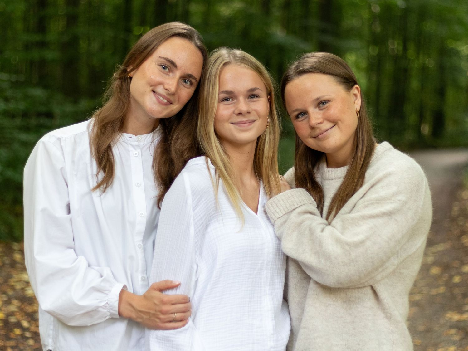 Three smiling young women in white and cream tops stand close together in a green forest setting.