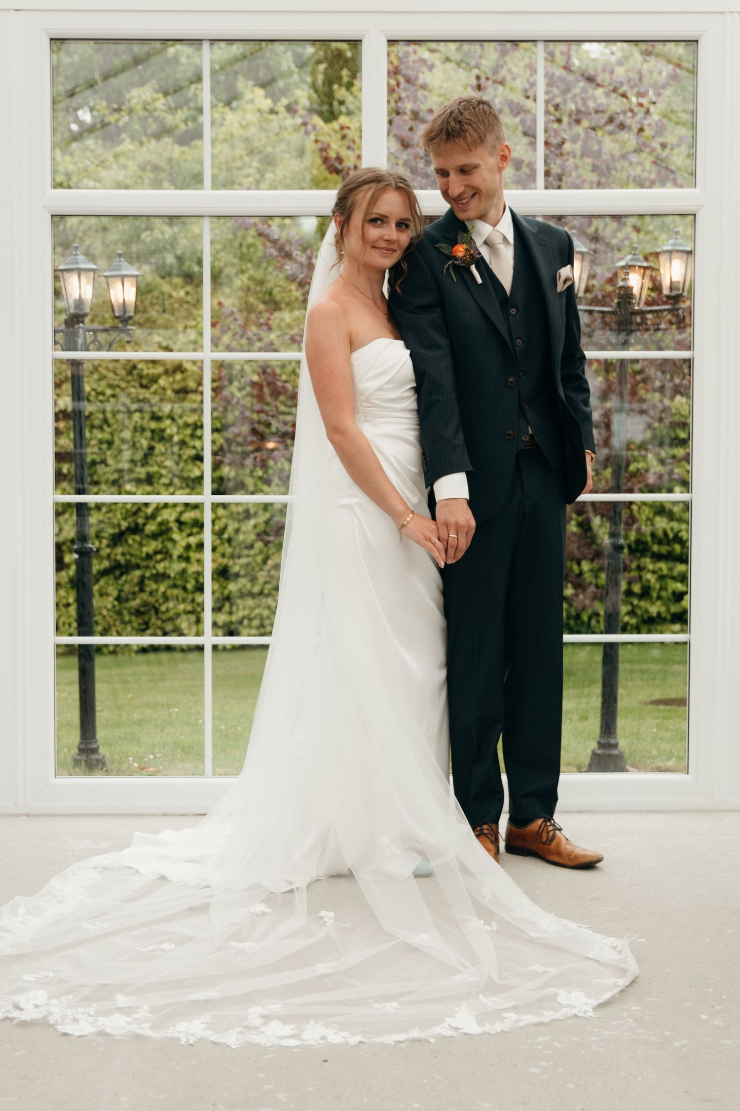 Groom embraces his bride in a strapless white gown with a long lace-trimmed veil in front of a large glass conservatory window.