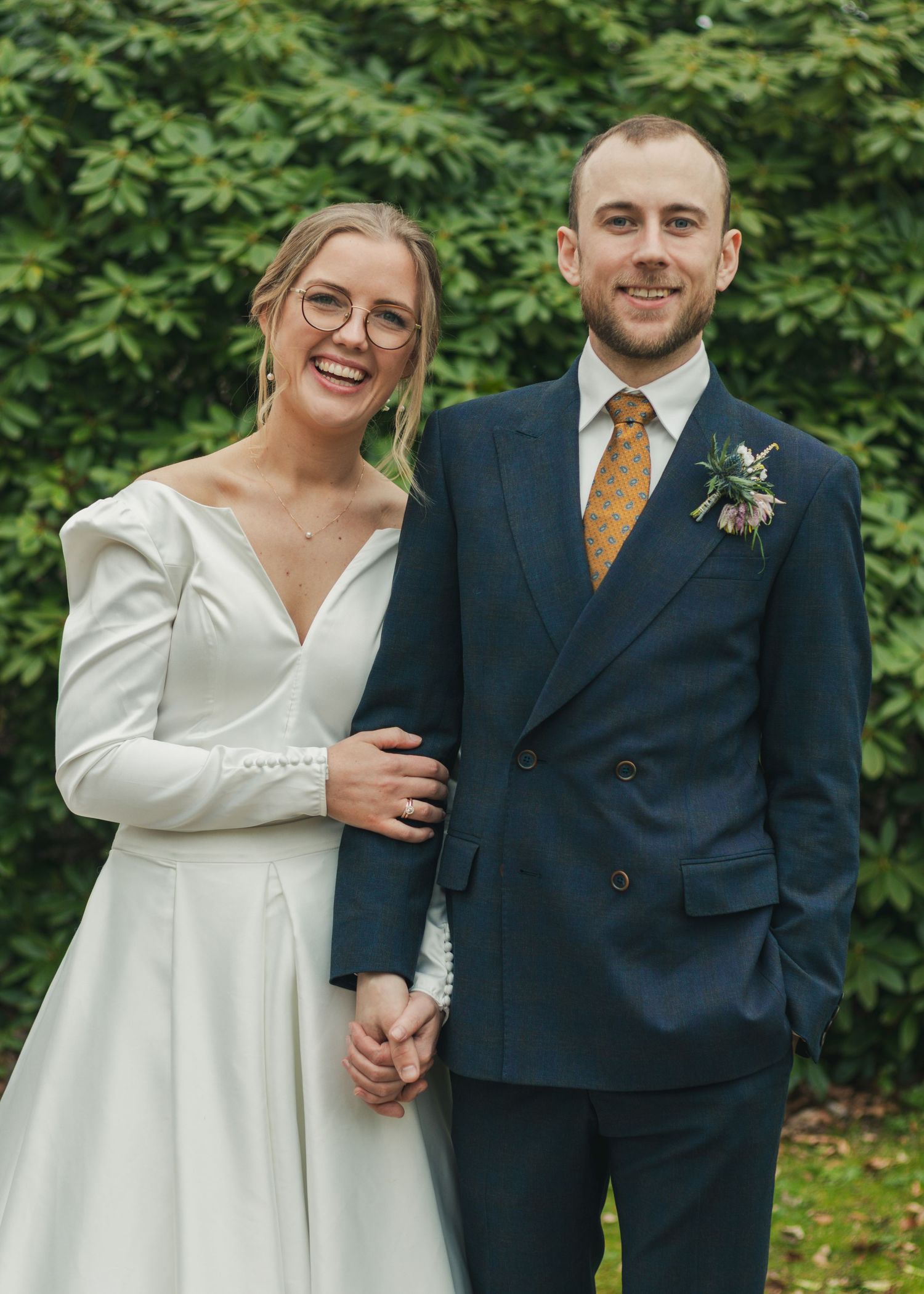 Happy newlyweds hold hands outdoors, the bride in a white gown with puffed sleeves and the groom in a navy double-breasted suit.