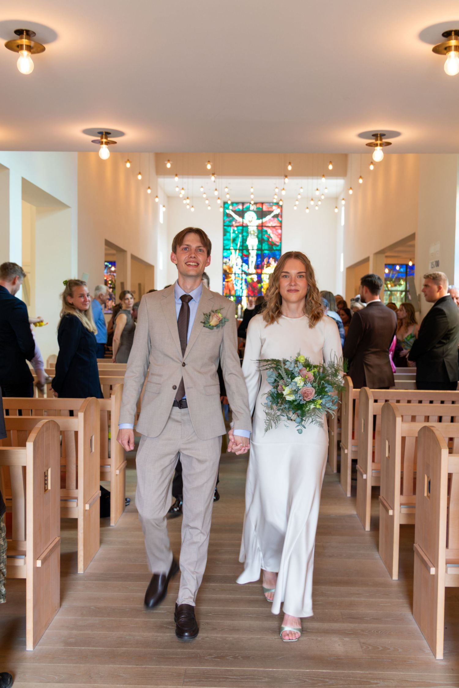 Bride with a green and pink bouquet and groom in a light suit walk hand in hand up a church aisle lined with wooden pews and guests.