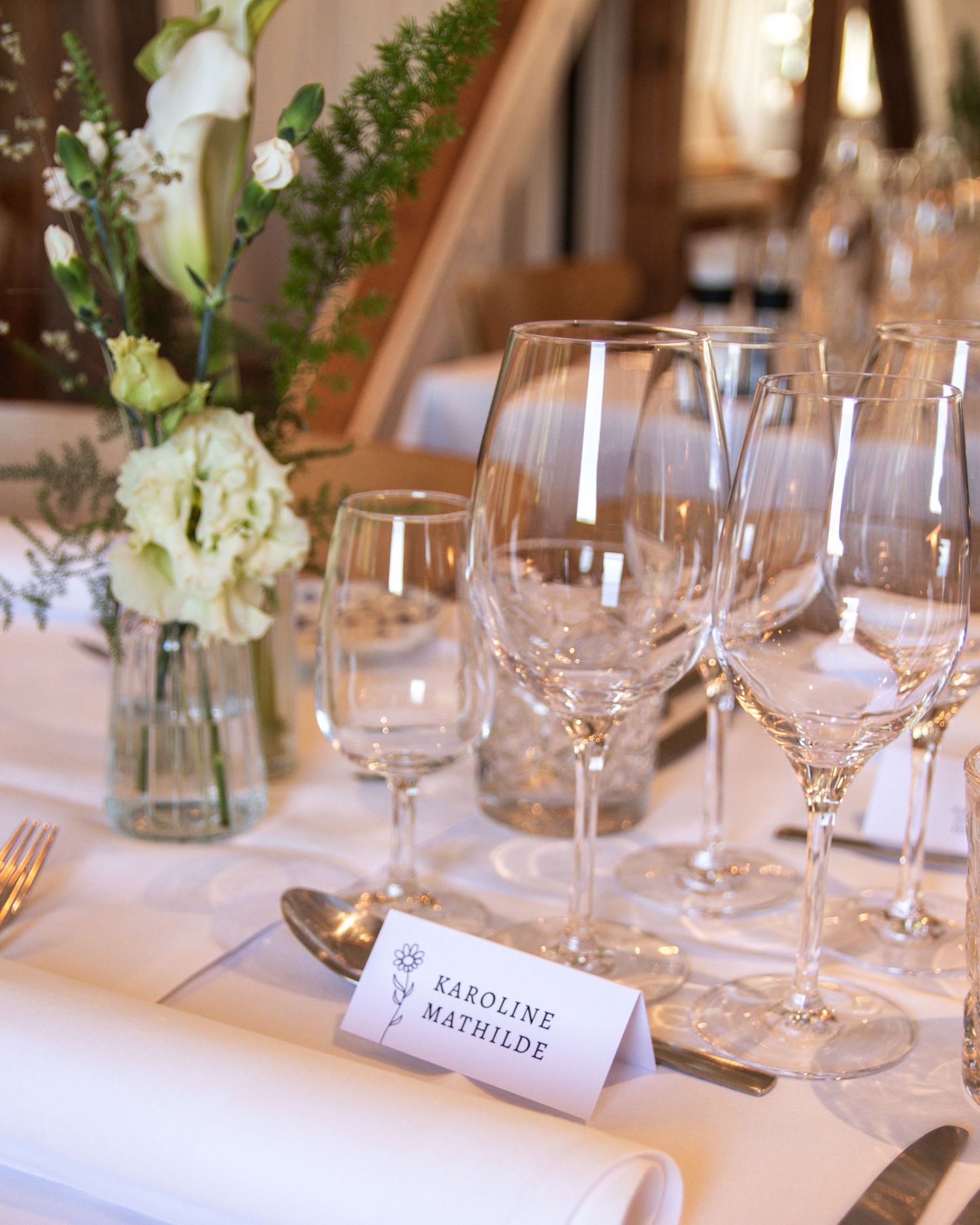 Elegant wedding table setting with crystal wine glasses, white linen, a flower vase and a place card reading Karoline Mathilde.