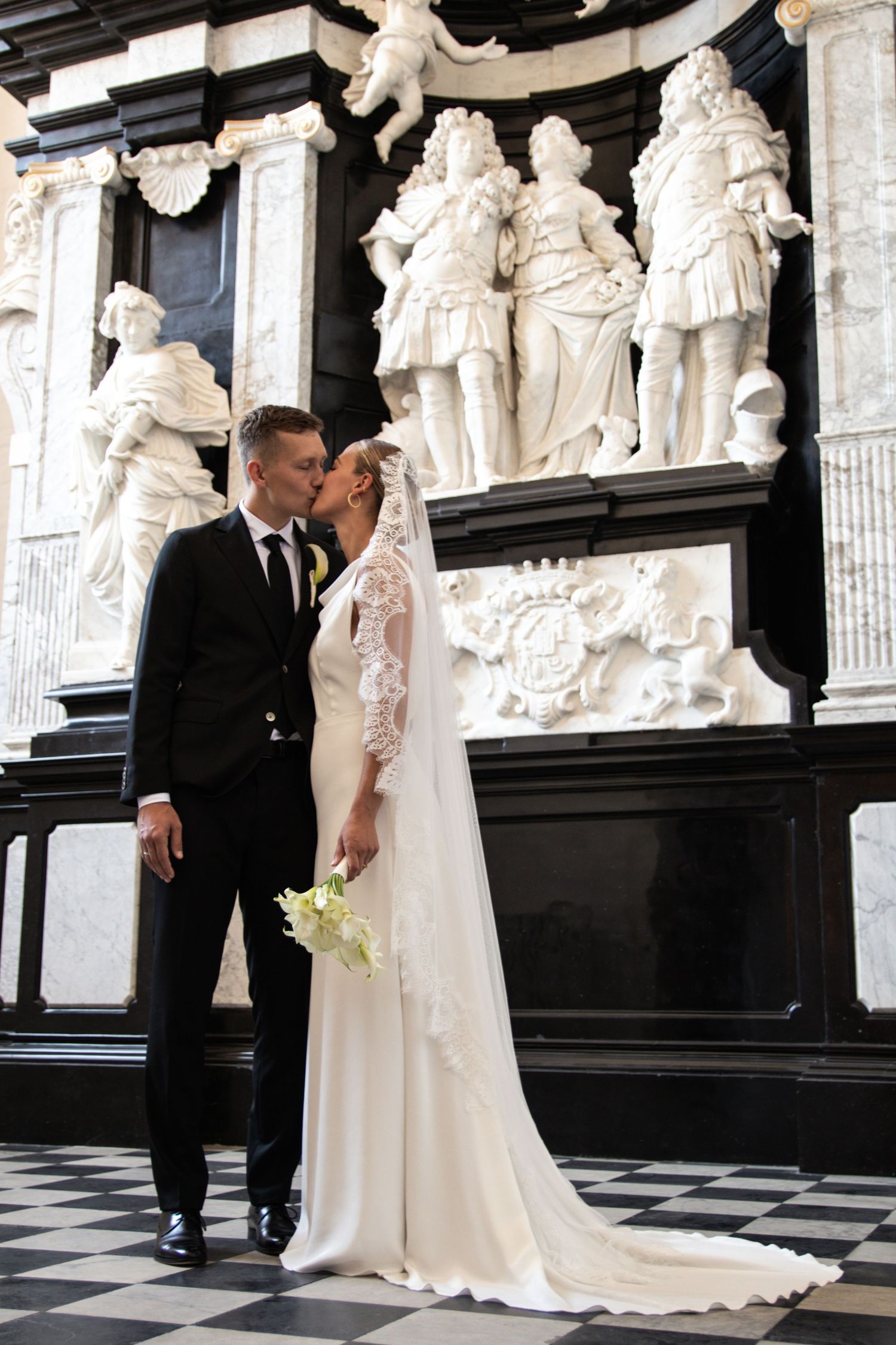 A bride and groom share a kiss in an ornate church before white marble statues and a black stone monument.