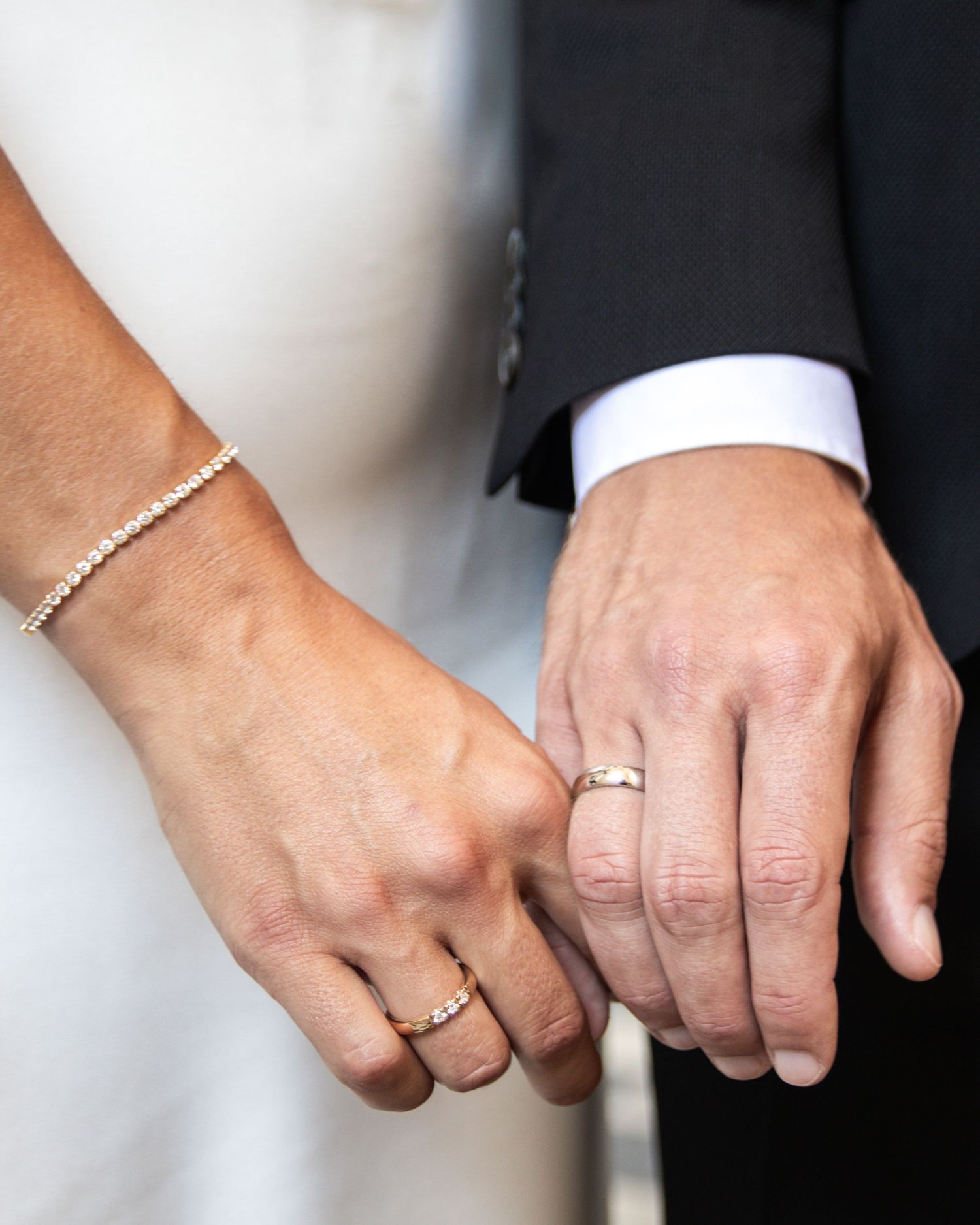 Close-up of a bride's and groom's clasped hands showing gold wedding rings and a sparkling bracelet against a white dress and dark suit.