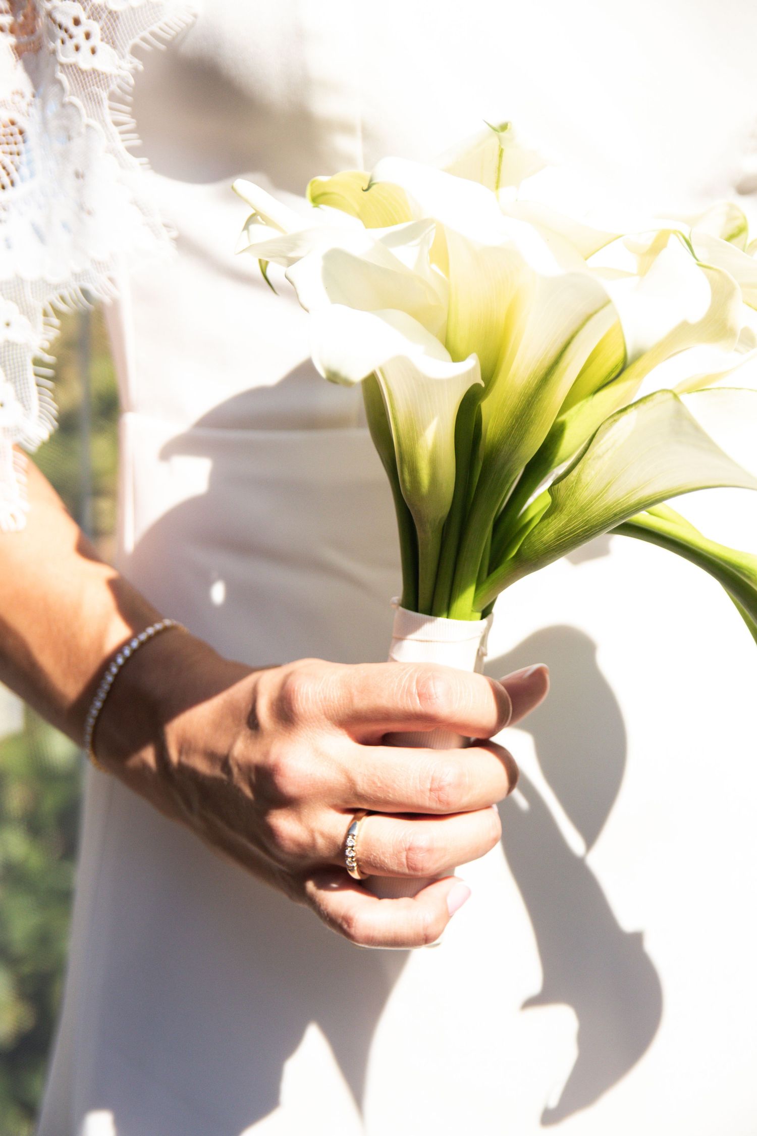 Bride in a white dress holds a bouquet of white calla lilies, showing her ring and a beaded bracelet.