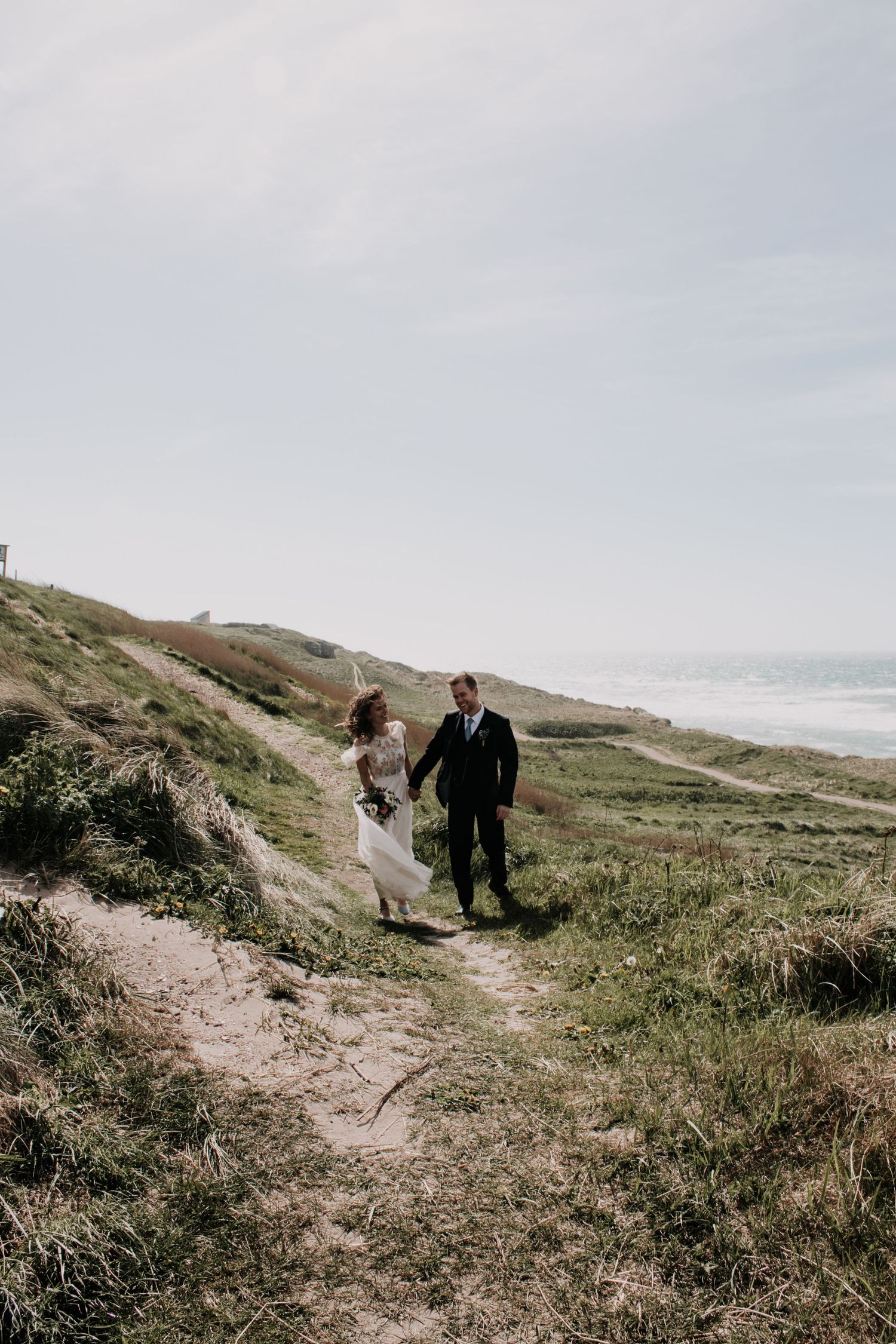 Bride in a flowing white dress and groom in a dark suit hold hands while walking along a grassy coastal dune path by the sea.