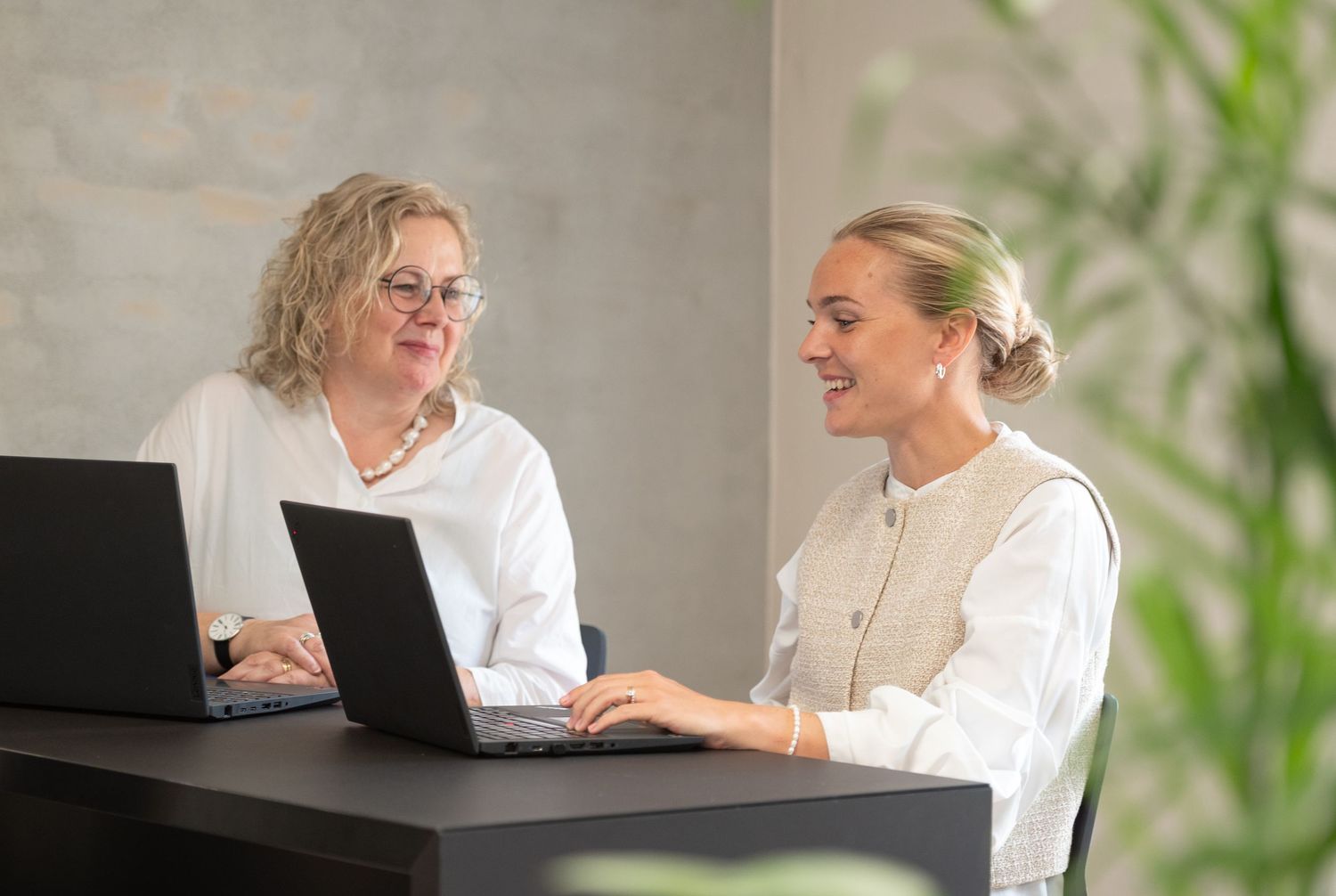 Two women in light clothing work together at a dark table with laptops, smiling as they discuss work in a modern office.