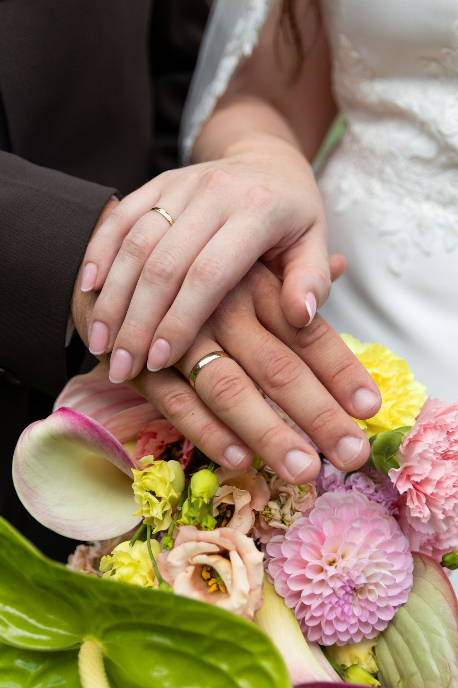 Close-up of a bride and groom's hands wearing wedding rings, resting on a bouquet of pink, yellow and cream flowers.